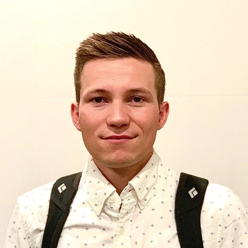 A man wearing a white patterned shirt and a black backpack, posing against a plain background.