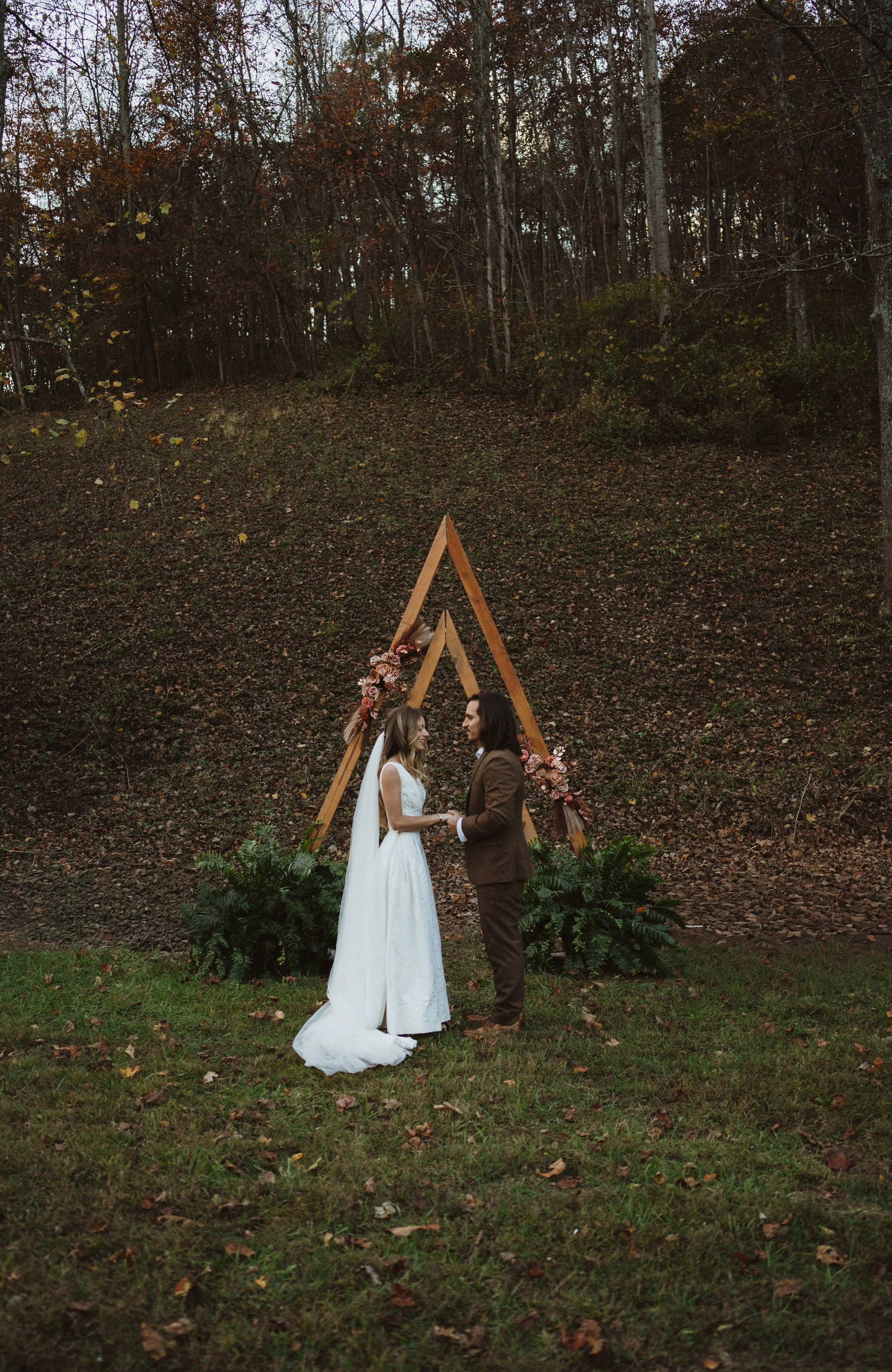 A bride and groom standing face to face holding hands during their outdoor wedding ceremony, with a triangular wooden arch decorated with pink and burgundy flowers behind them, set against a wooded hillside.