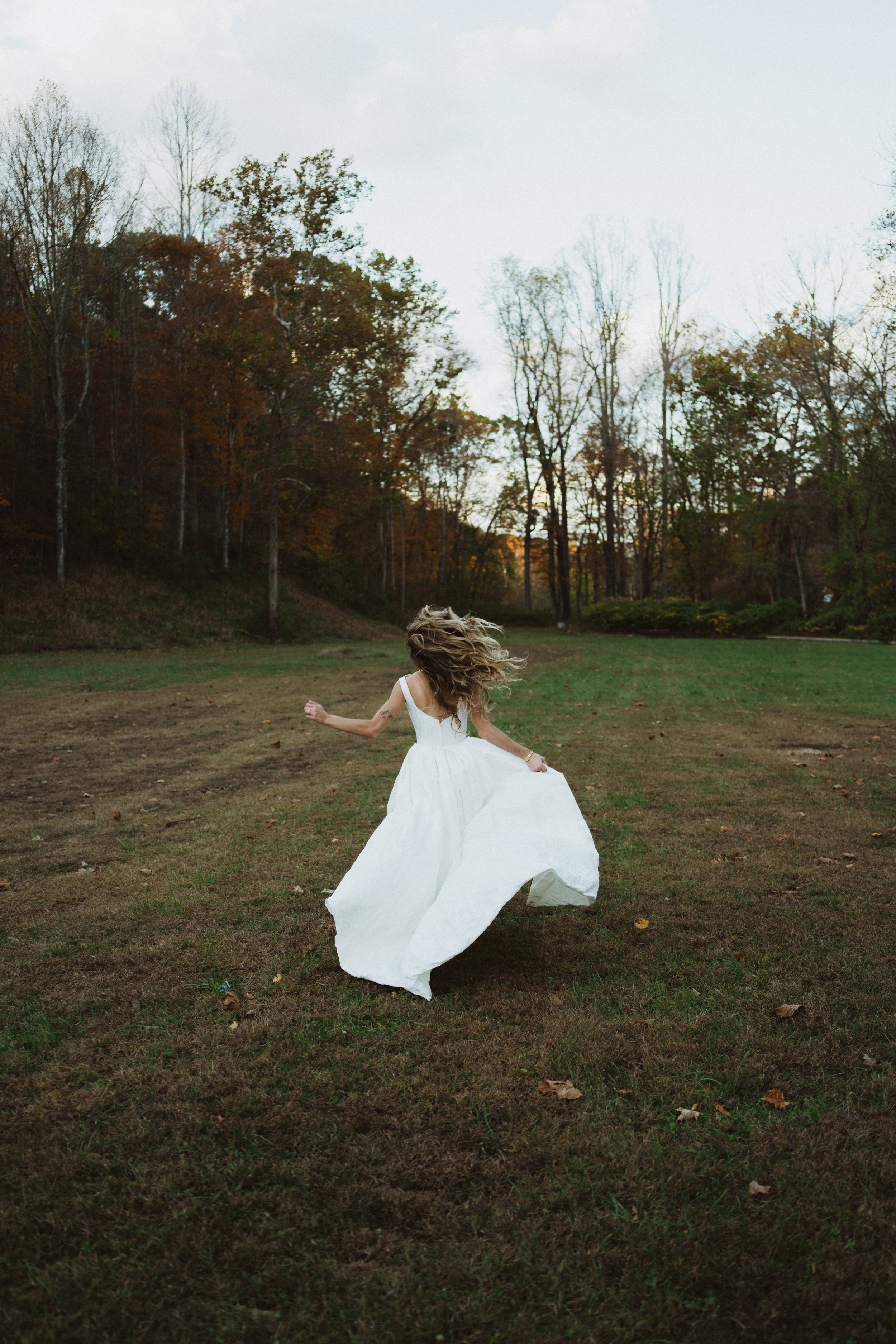 A woman in a white dress spinning in an open grassy field with trees in the background under a cloudy sky.
