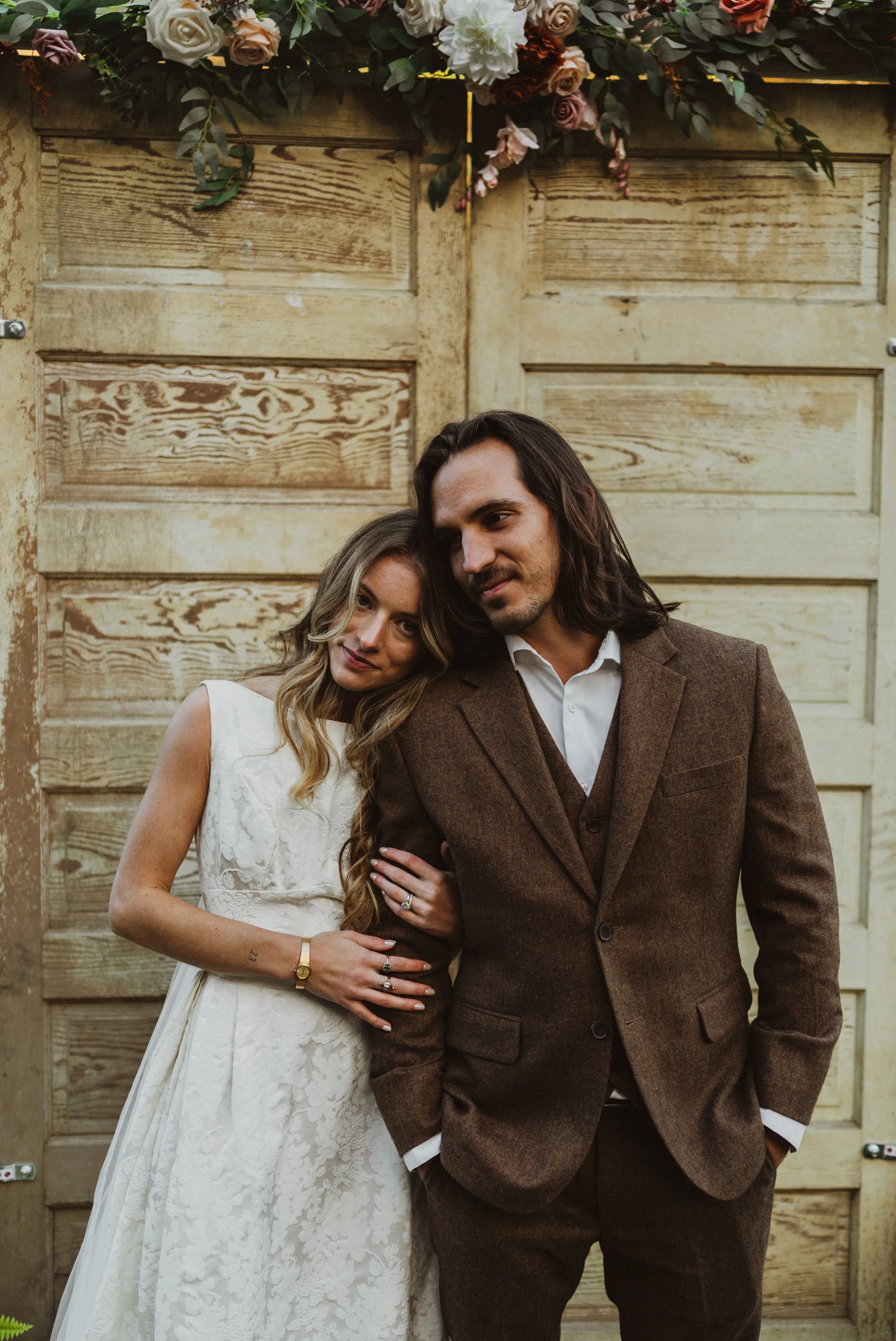 A couple stands close together in front of a rustic wooden background adorned with a floral arrangement at the top. The woman wears a white lace dress and the man wears a brown suit.