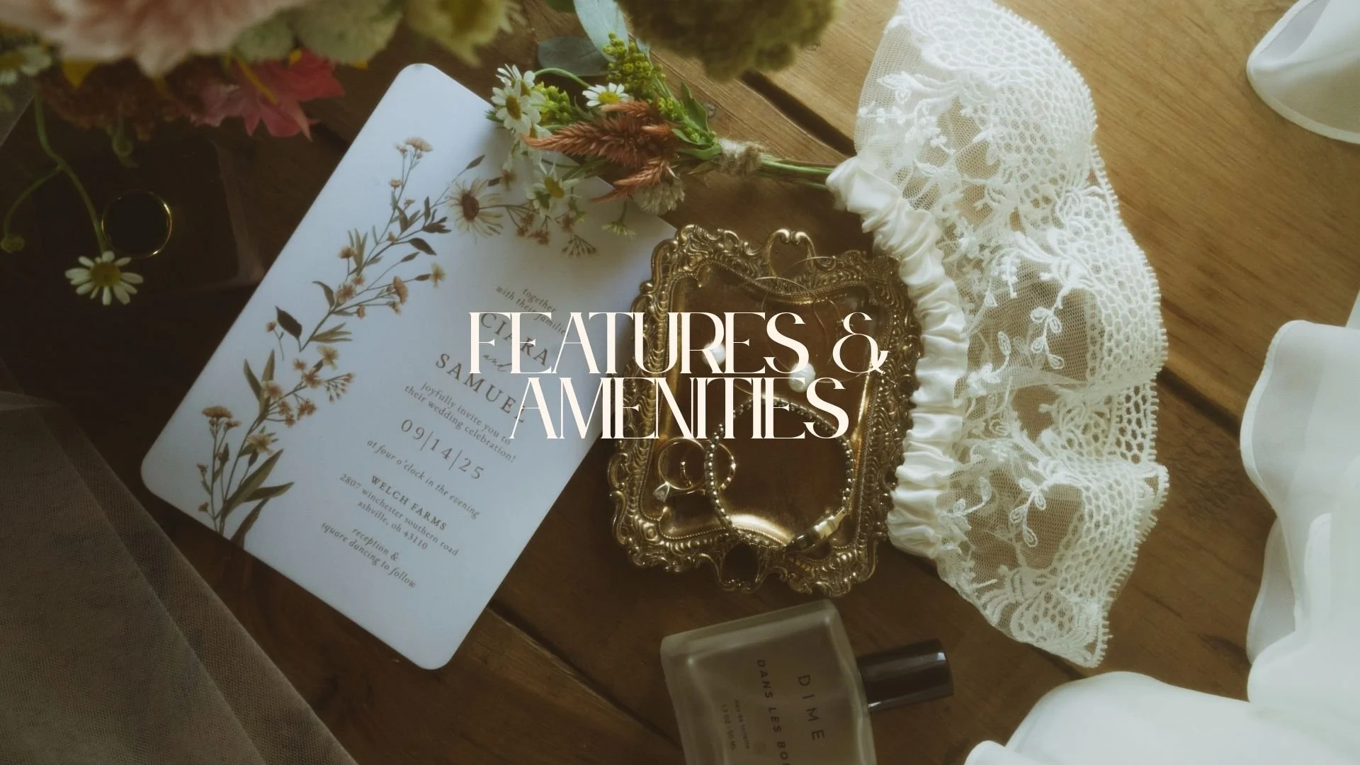 A wedding invitation card, a small bouquet of flowers, a vintage gold tray with jewelry, a bottle of perfume, and a lace fabric are arranged on a wooden surface.