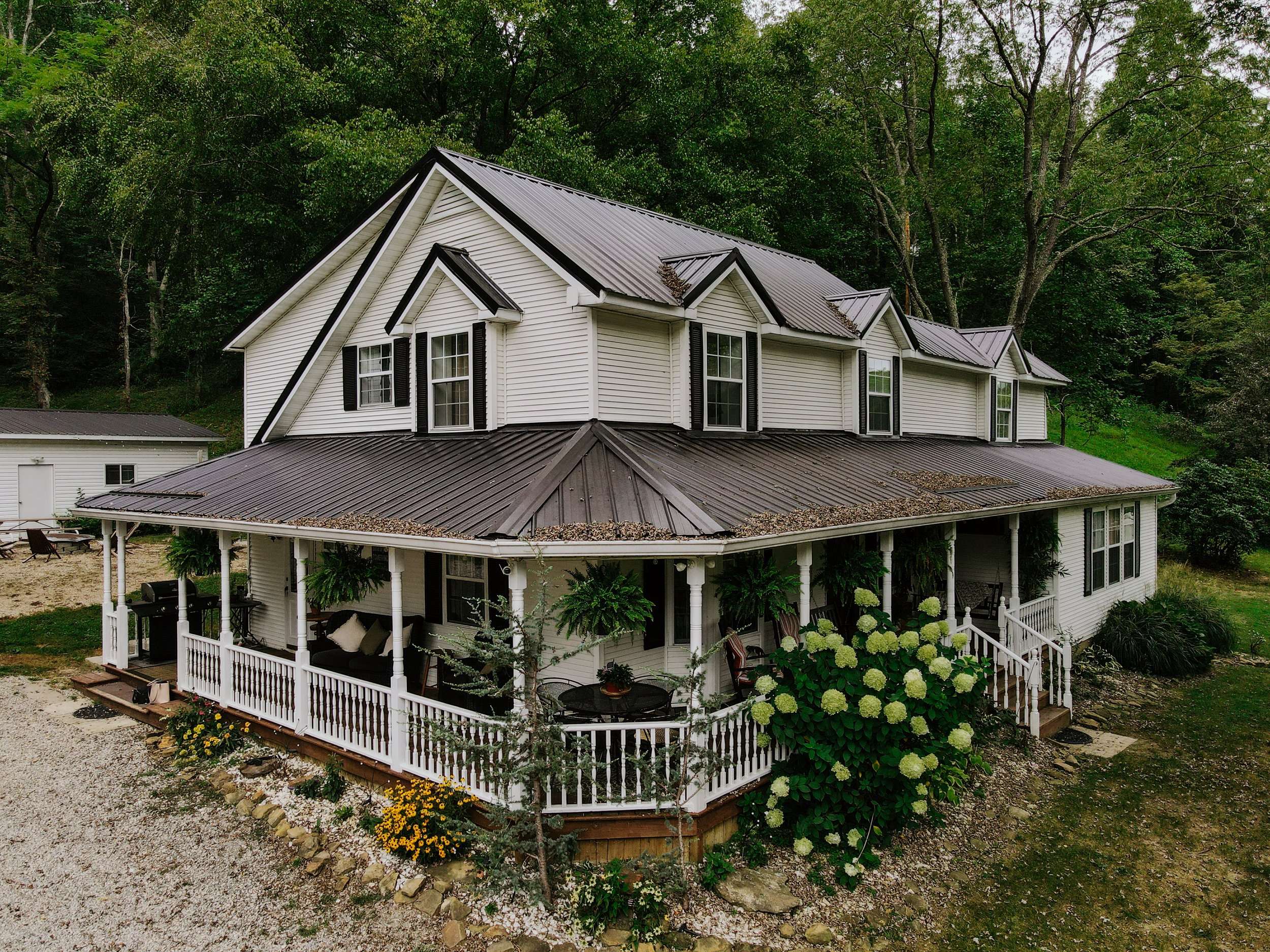 A two-story white house with a metal roof and a wraparound porch surrounded by green trees and garden plants.