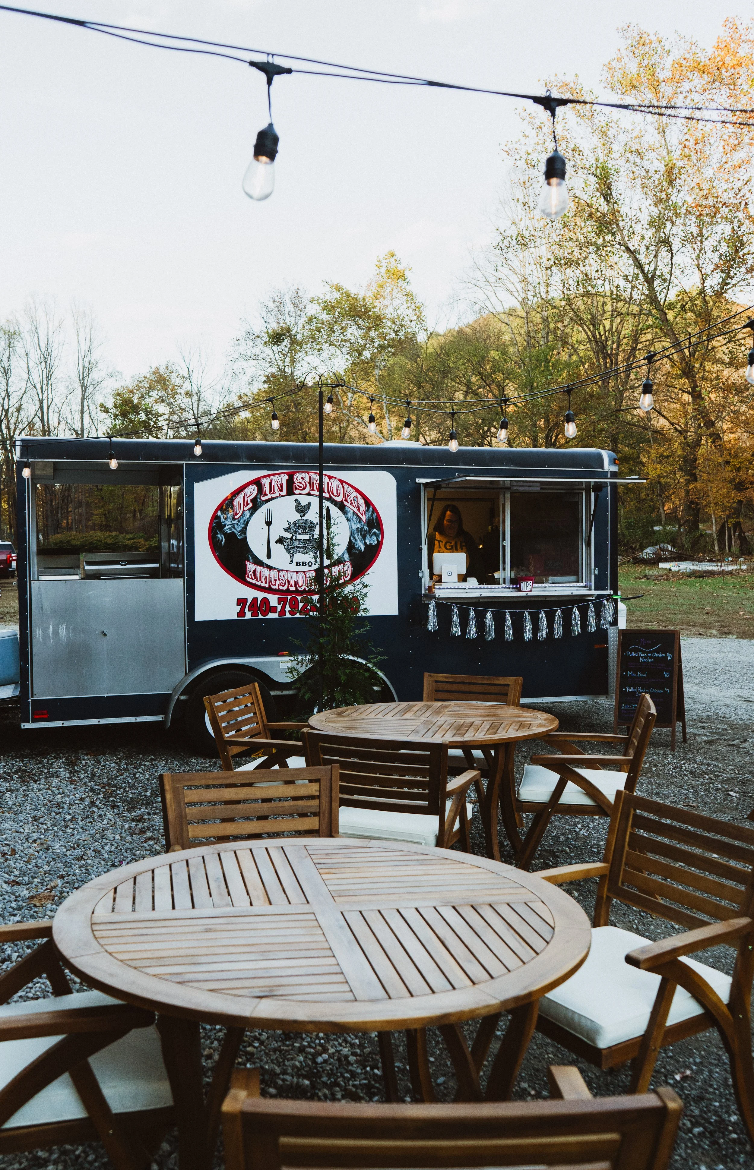 Outdoor scene with a BBQ food truck named "Up in Smoke" serving food, surrounded by wooden tables and chairs, with string lights overhead and trees with autumn foliage in the background.