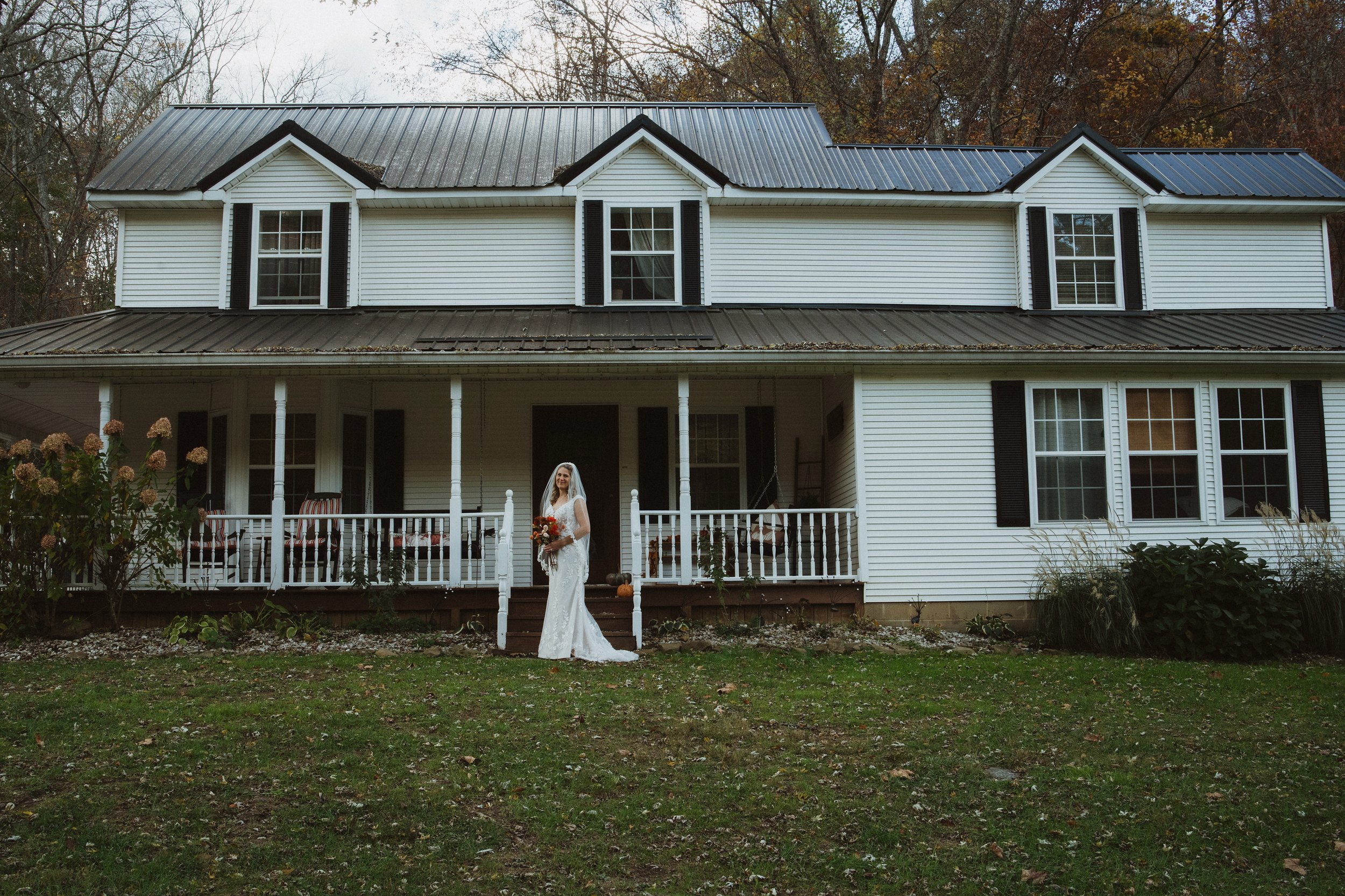A bride in a white wedding dress holding a bouquet standing on the front porch of a large white house with black shutters and a metal roof, surrounded by trees and grass.