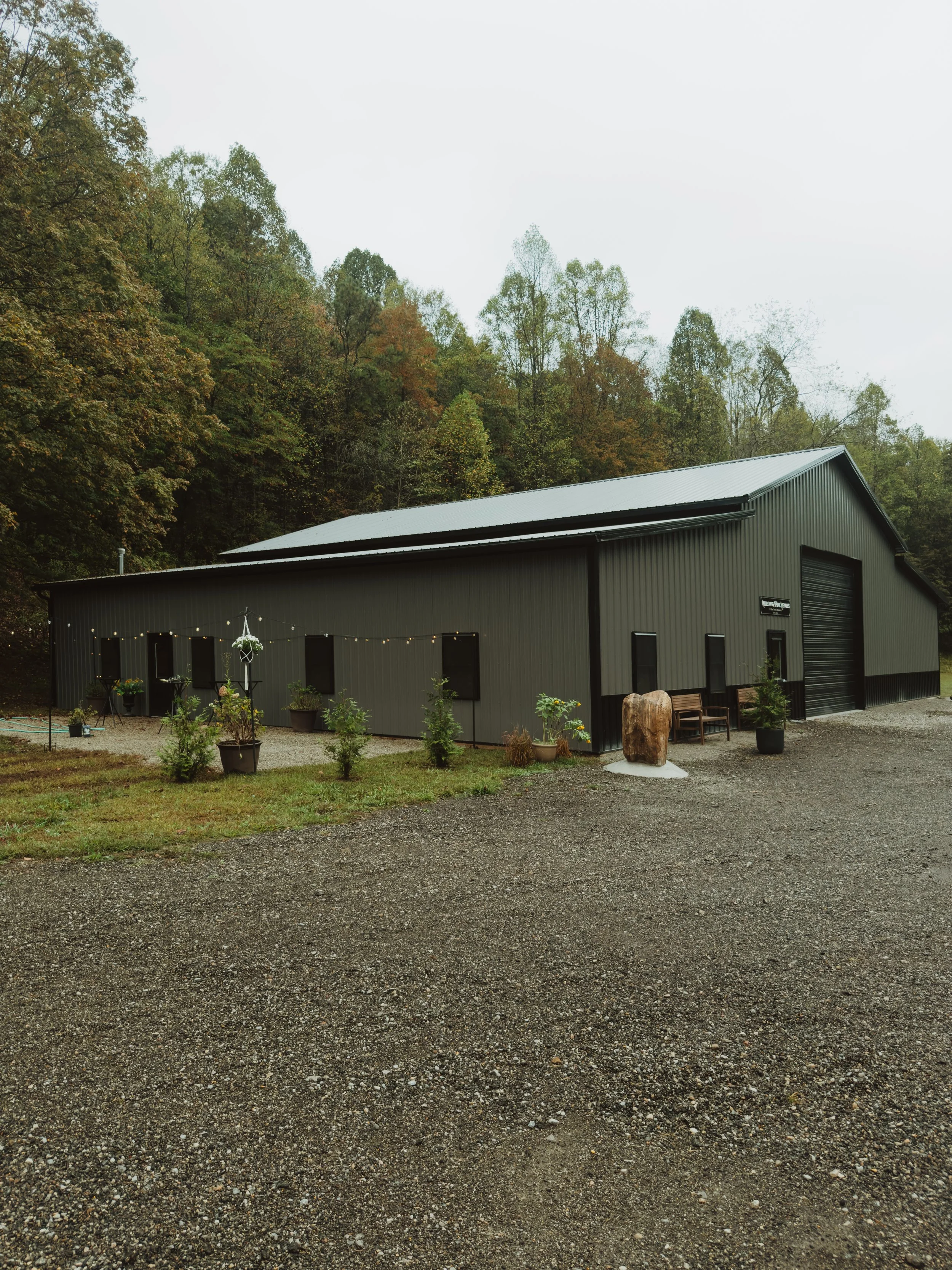 A large gray metal building with a sloped roof, surrounded by trees and small plants, with a gravel driveway in front.