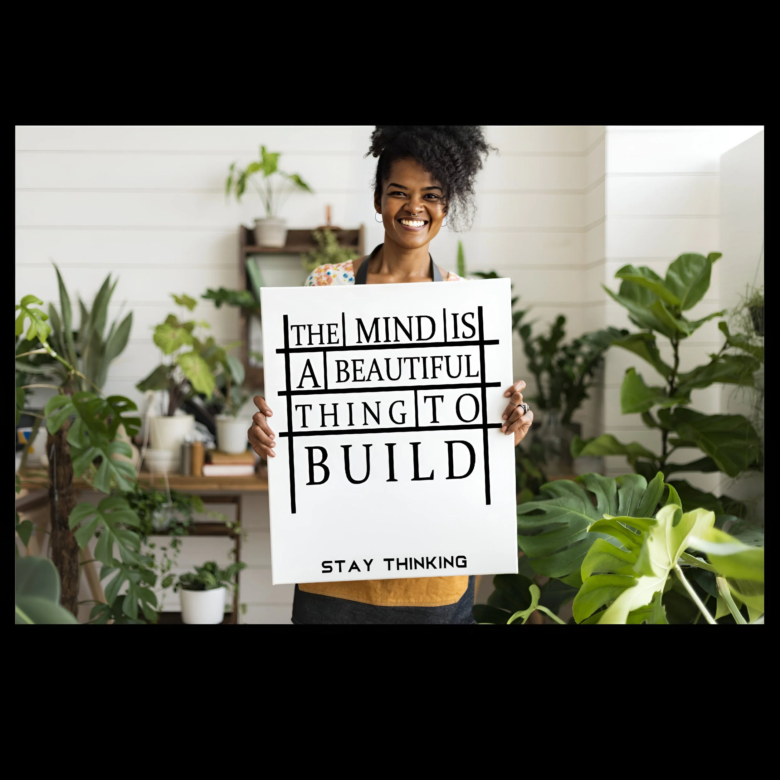 A woman holding a sign with the quote, "The mind is a beautiful thing to build. Stay thinking," in a room with numerous green plants and white walls.