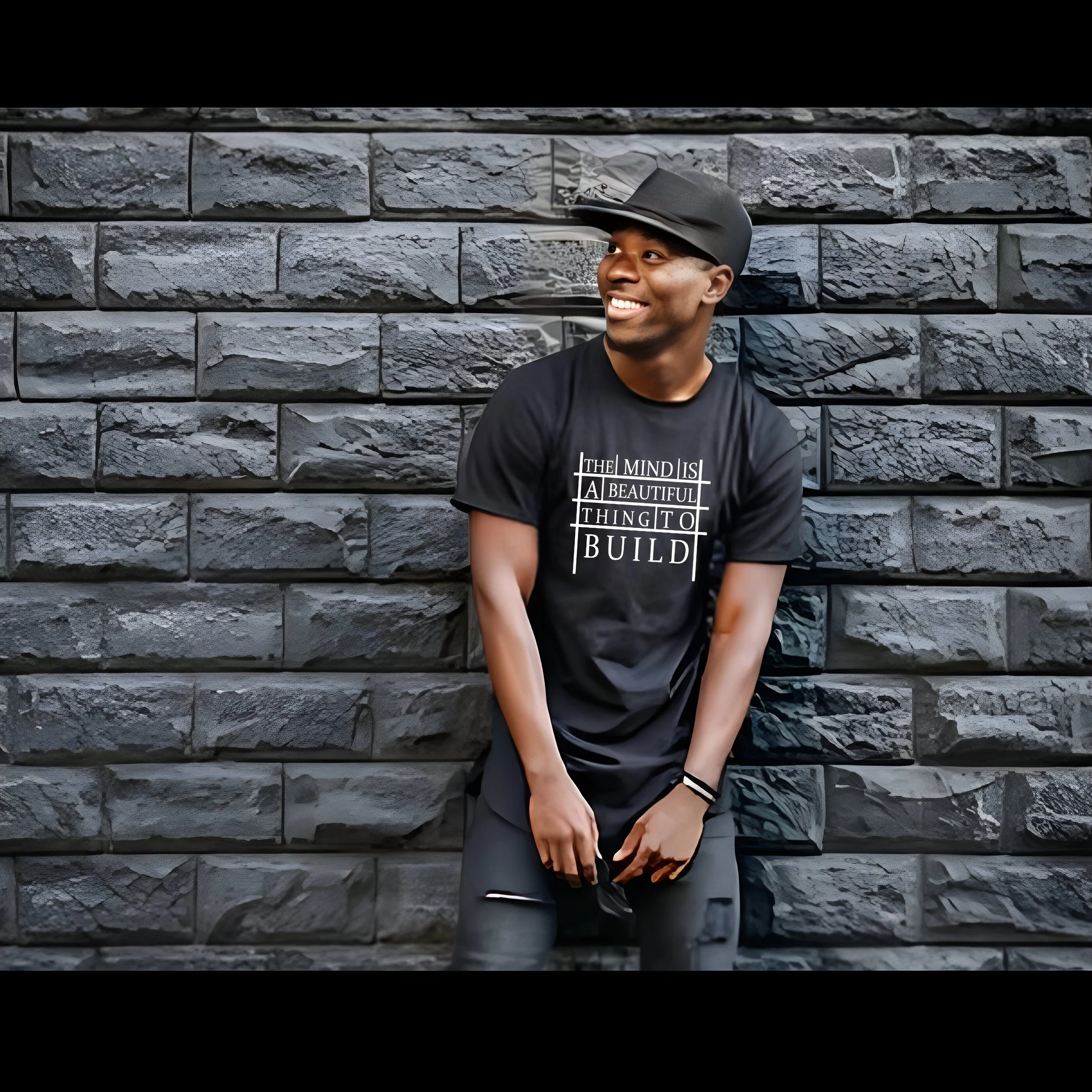 Young man smiling, wearing a black hat and a black T-shirt with a quote, standing against a textured gray stone wall.