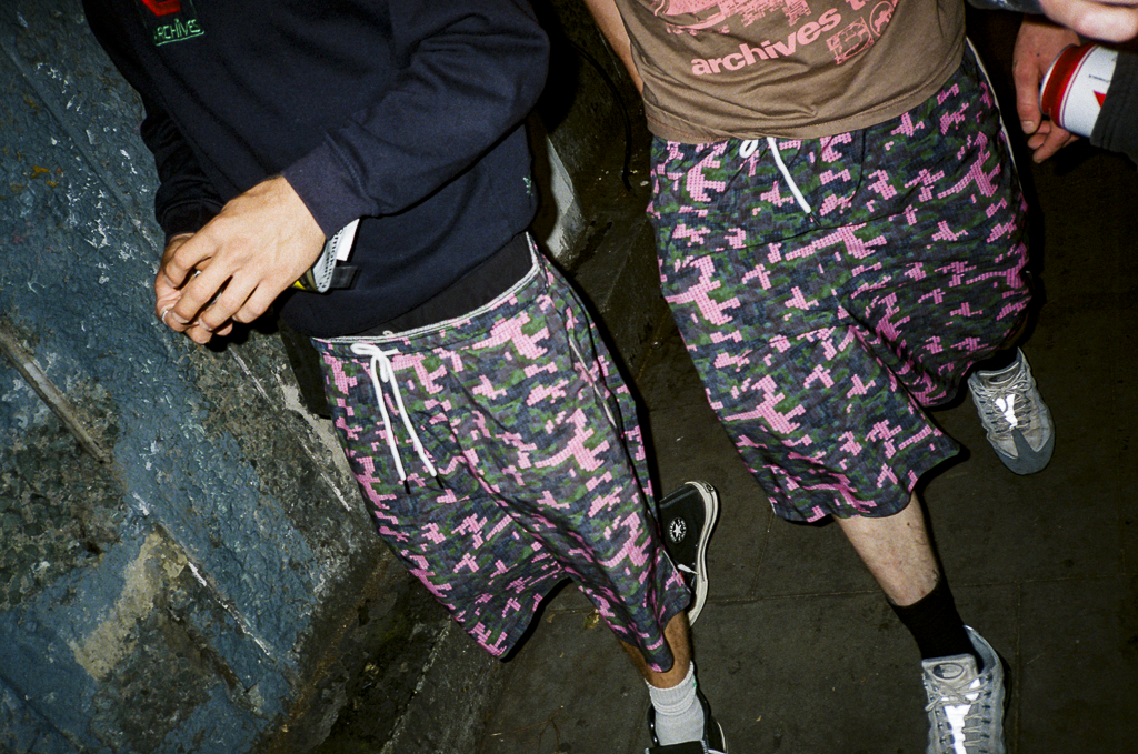 Two people wearing shorts with a pink and black camouflage pattern, standing indoors near a blue brick wall. One person is wearing a black jacket and the other is wearing a brown T-shirt.