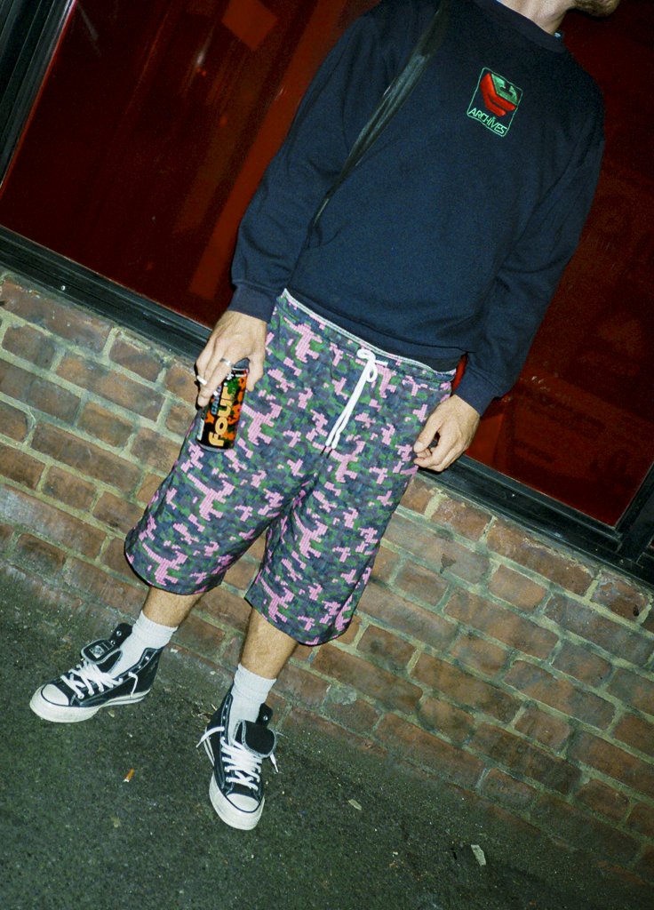 A person standing outside wearing a black long-sleeve shirt, pink and green camo shorts, white socks, and black and white sneakers. The person is holding a beverage can in their right hand.