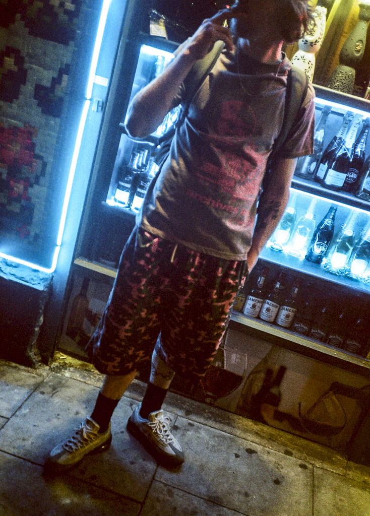 A person standing in front of a glass display with alcohol bottles, making a peace sign with one hand and holding their phone to their face with the other, wearing a graphic t-shirt, camouflage shorts, and sneakers.
