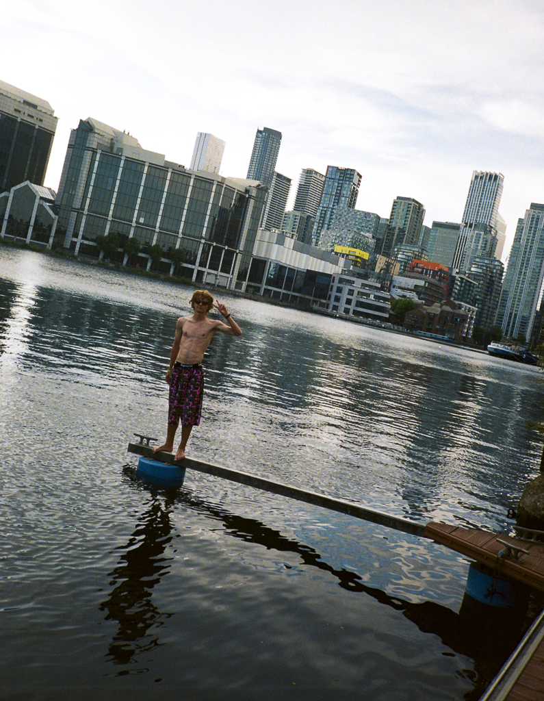 A shirtless man with sunglasses and patterned shorts standing on a small floating platform on a body of water with a city skyline in the background.