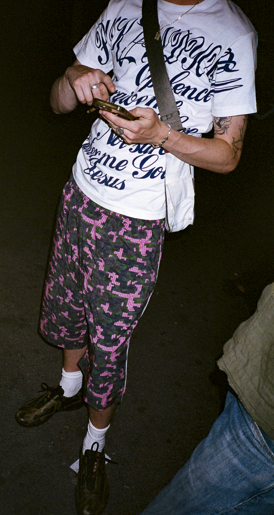 Person wearing a white T-shirt with blue script and camouflage shorts with pink and purple patterns, holding a phone, standing on a dark ground.