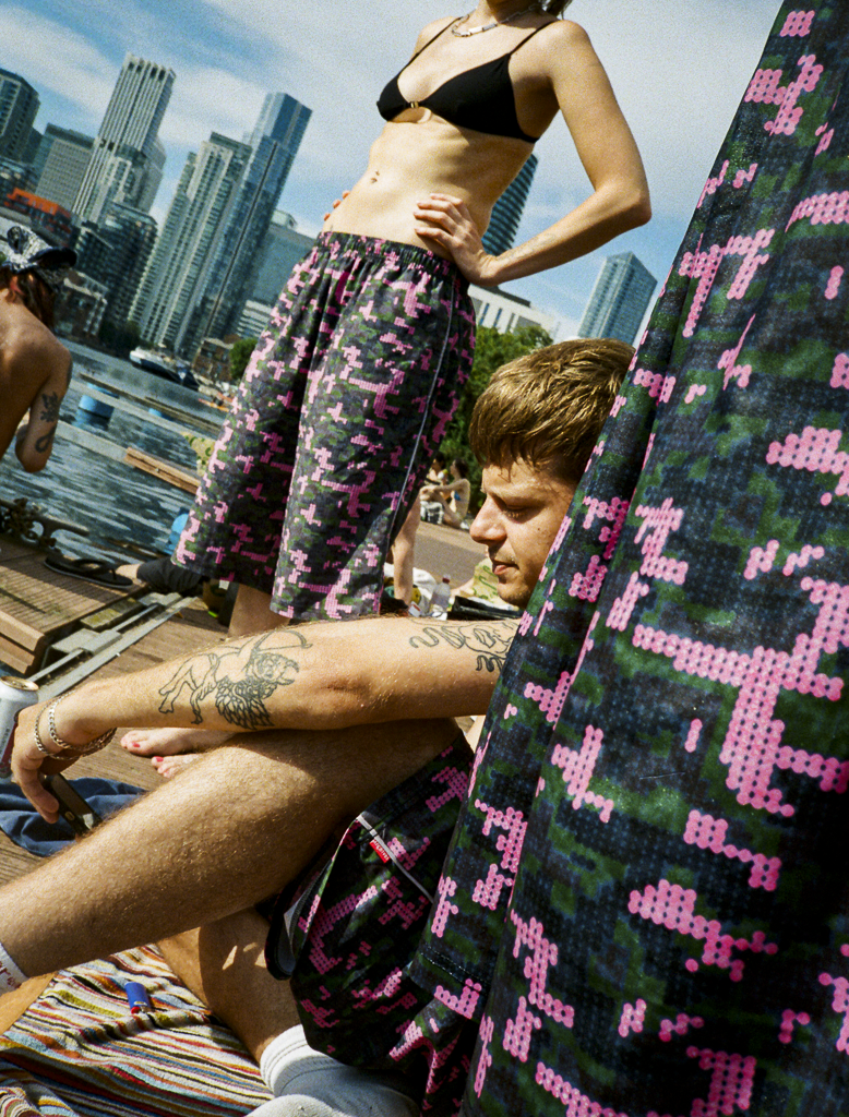 A woman in a black bikini top and colorful patterned pants standing with her hand on her hip, and a man with tattoos sitting outdoors near a city skyline, with colorful patterned fabric in the foreground.