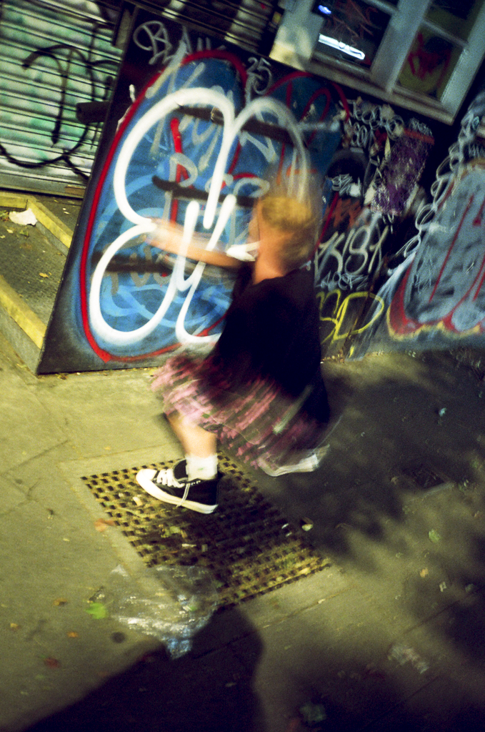 A young girl with blonde hair wearing a black shirt, a pink plaid skirt, and black sneakers with white soles and laces, spray-painting graffiti art on a street billboard at night surrounded by other graffiti. She is facing the billboard, with her arm extended to spray paint.