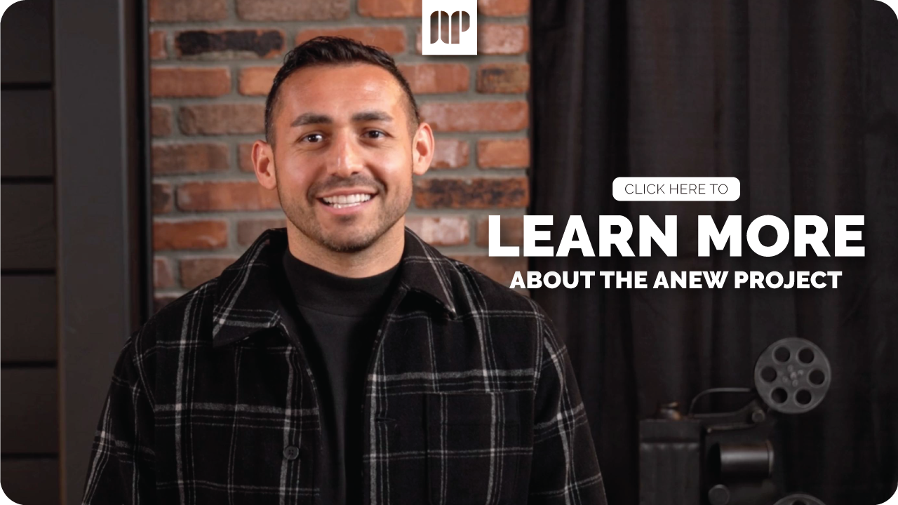 Smiling man in black plaid shirt with brick wall background and black curtain, promoting the Anew project.