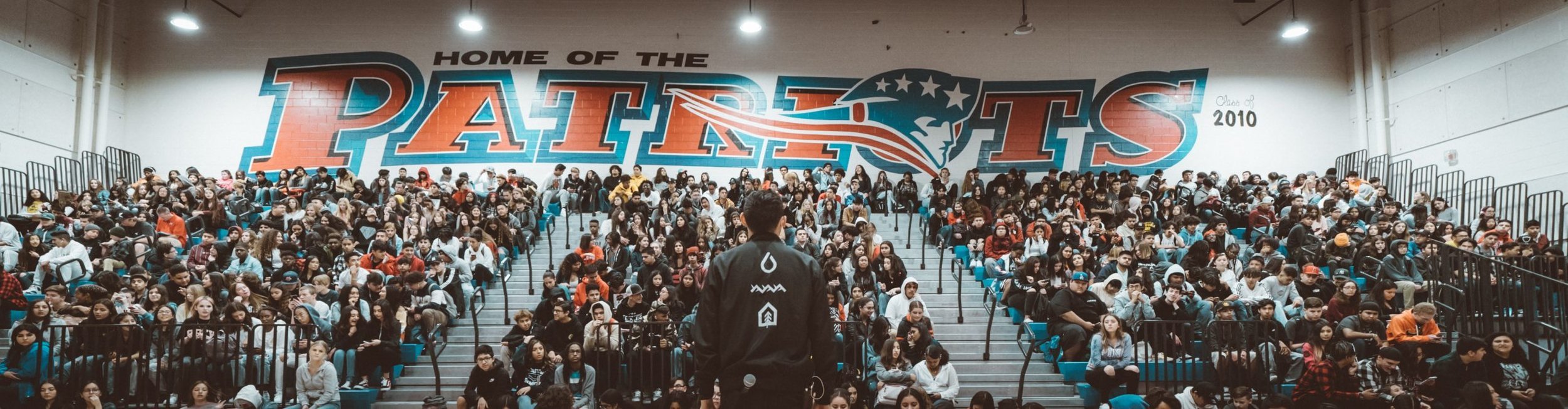 A large indoor gymnasium filled with students seated in bleachers, with a man standing in front of them holding a microphone. The wall behind has a large mural reading 'Home of the Patriots' with patriotic elements and the year 2010.