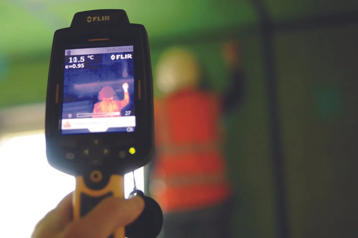Handheld FLIR thermal imaging camera displaying a thermal image of a person in an orange safety vest and hard hat, standing in a room with green walls, with a blurred background.