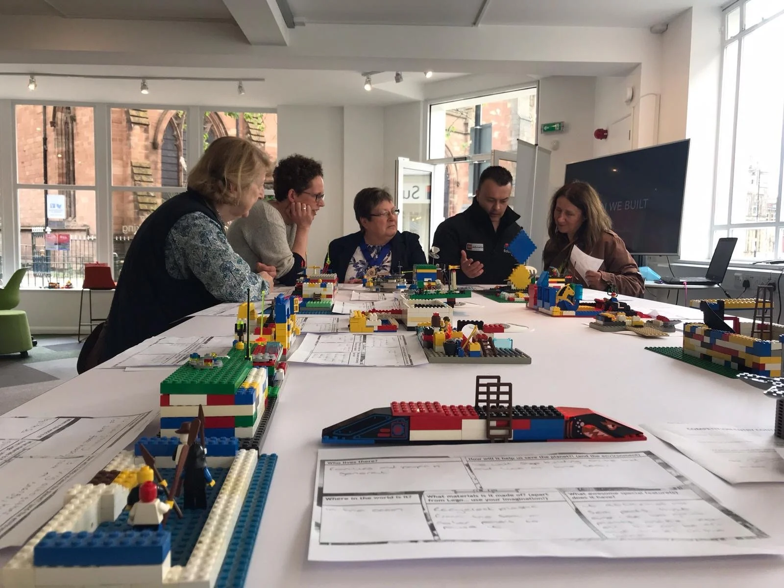 Six people sitting around a large table with various colorful LEGO structures and papers, inside a bright room with large windows.