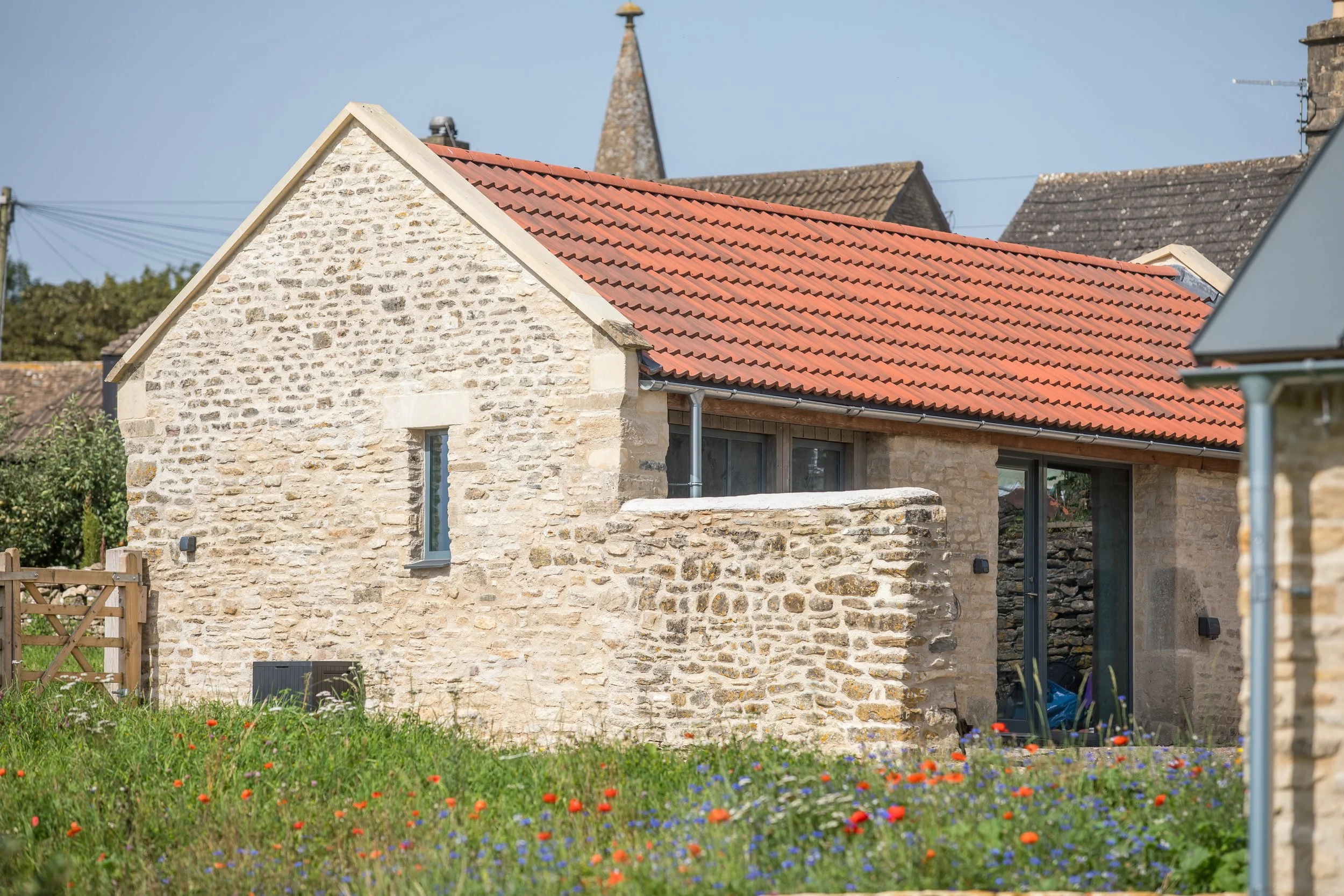 A stone house with a red tiled roof and a small balcony, surrounded by a grassy garden with red and purple flowers.