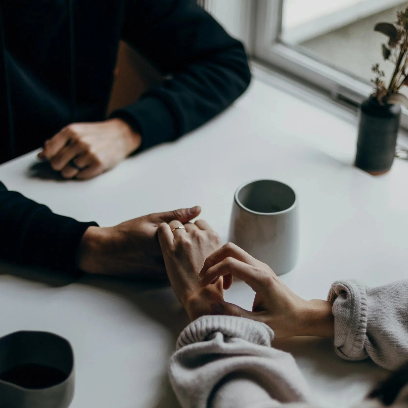 Two people holding hands on a white table near a window, with coffee cups and a potted plant visible.