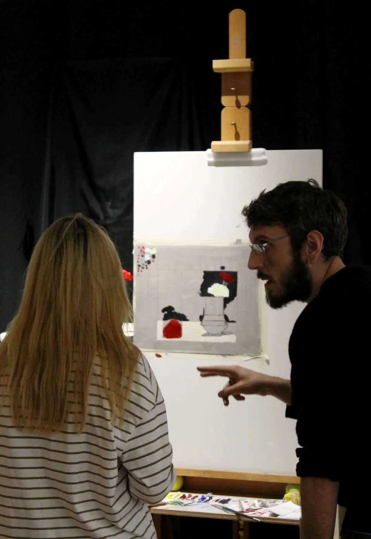 Two people are standing and talking in front of an art display. The display includes a piece of art with a black silhouette of a bird, a red flower, and other decorative elements. Behind the art, there is a white wall or panel, and above it, a wooden