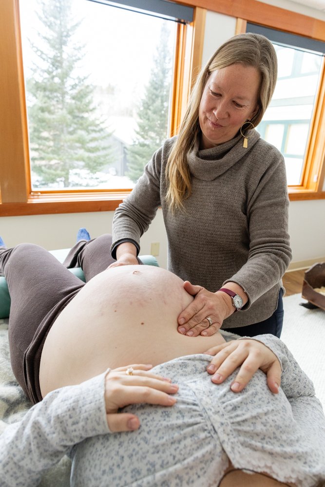 A woman is practicing pregnancy massage on a pregnant person lying on a massage table inside a room with large windows revealing a snowy landscape outside.