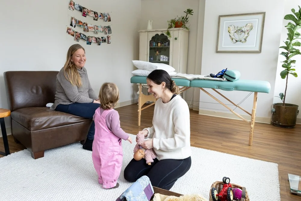 A woman kneeling on a white rug, handing a toy to a young girl in pink overalls, with an older woman sitting on a brown leather couch watching them and smiling, in a room with a massage table, framed artwork, and photos on the wall.