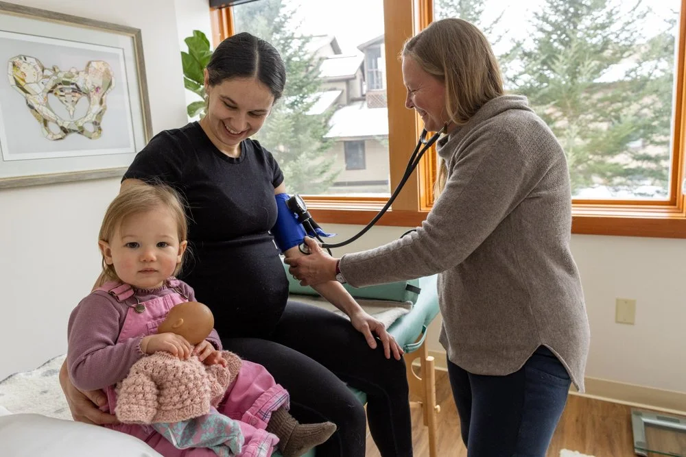 A pregnant woman sitting on an examination table in a medical room is having her blood pressure taken by a healthcare provider. A young girl sits beside her, holding a doll and looking at the camera. The room has large windows with outside trees and houses visible.