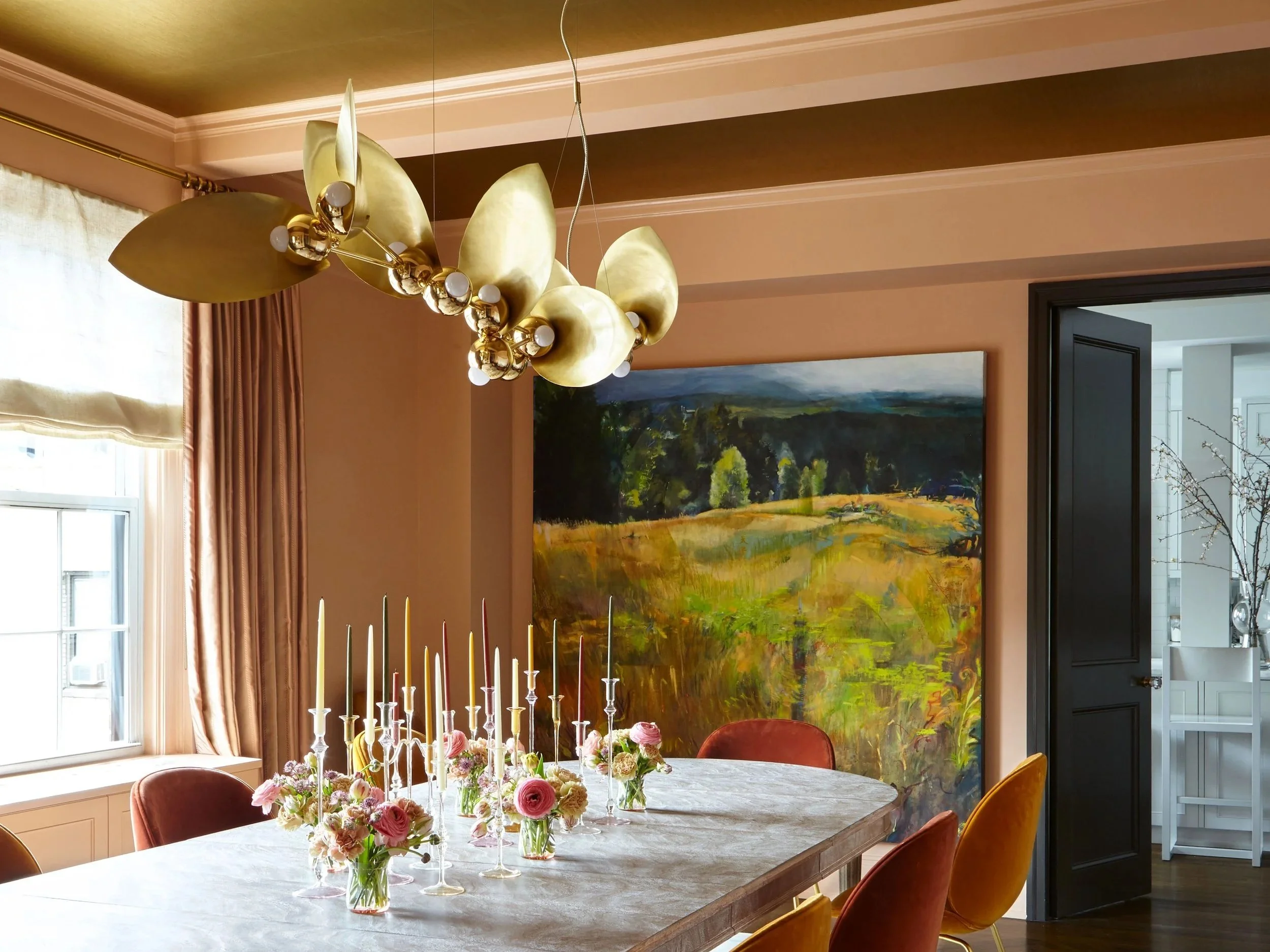 Large gold chandelier light hanging over modern table with taper candles, orange dining chairs, and modern landscape painting
