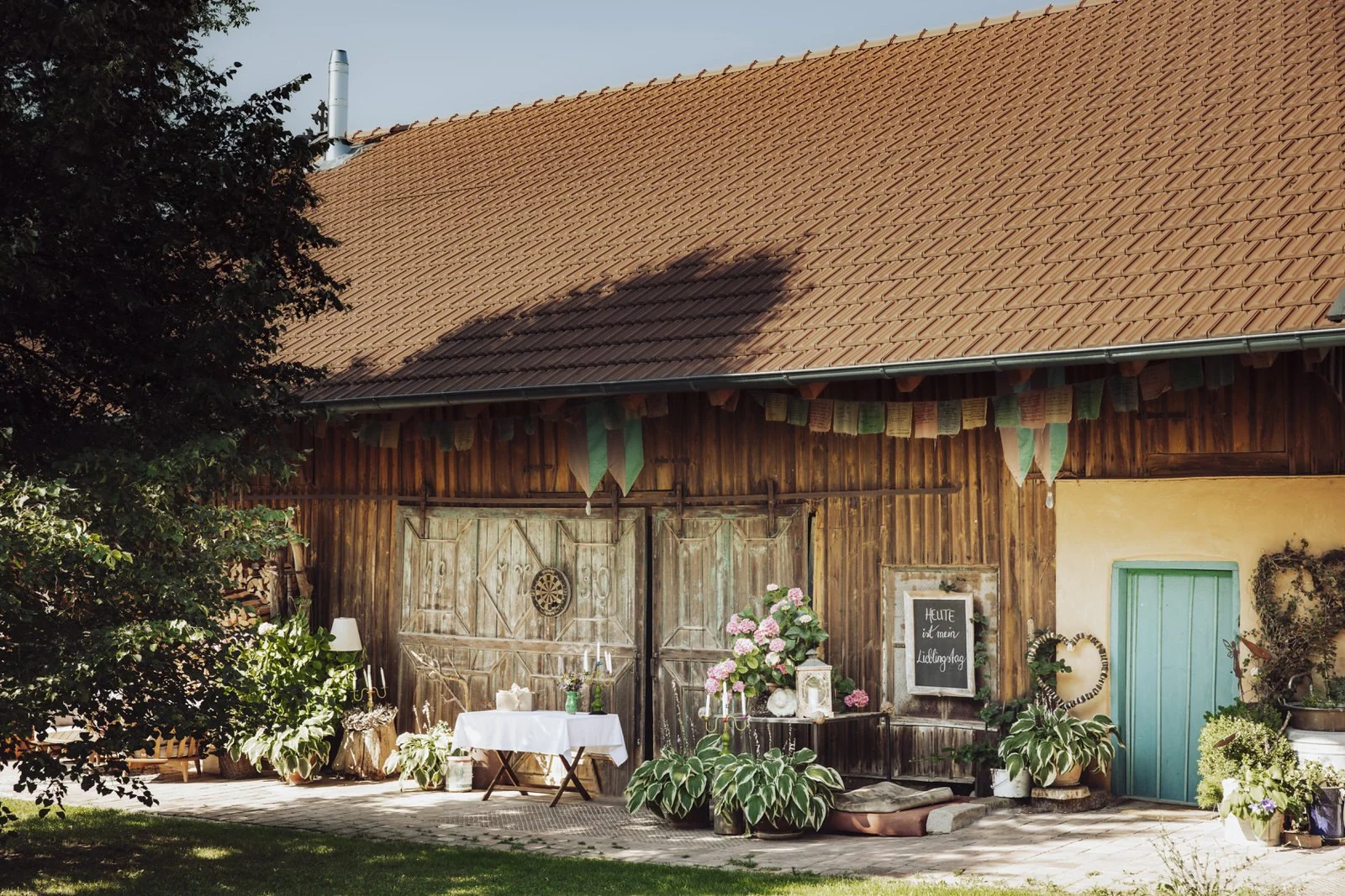 Ein Bauernhof mit Holzfassade, grün gestrichenem Tür und Blumenschmuck im Garten. Es gibt einen gedeckten Tisch mit Kerzen und Blumen, eine Tafel mit Beschriftung, und hängt Gebetsfahnen unter dem Dach. Ein Schatten eines Baumes fällt auf das Dach.