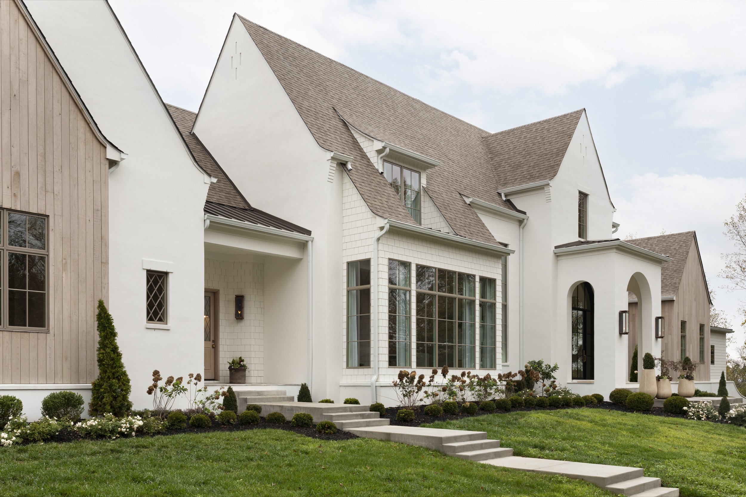 A modern house with a combination of white stucco and light wood siding, multiple gabled roofs, and large windows, surrounded by a manicured lawn and garden with shrubs and small plants.