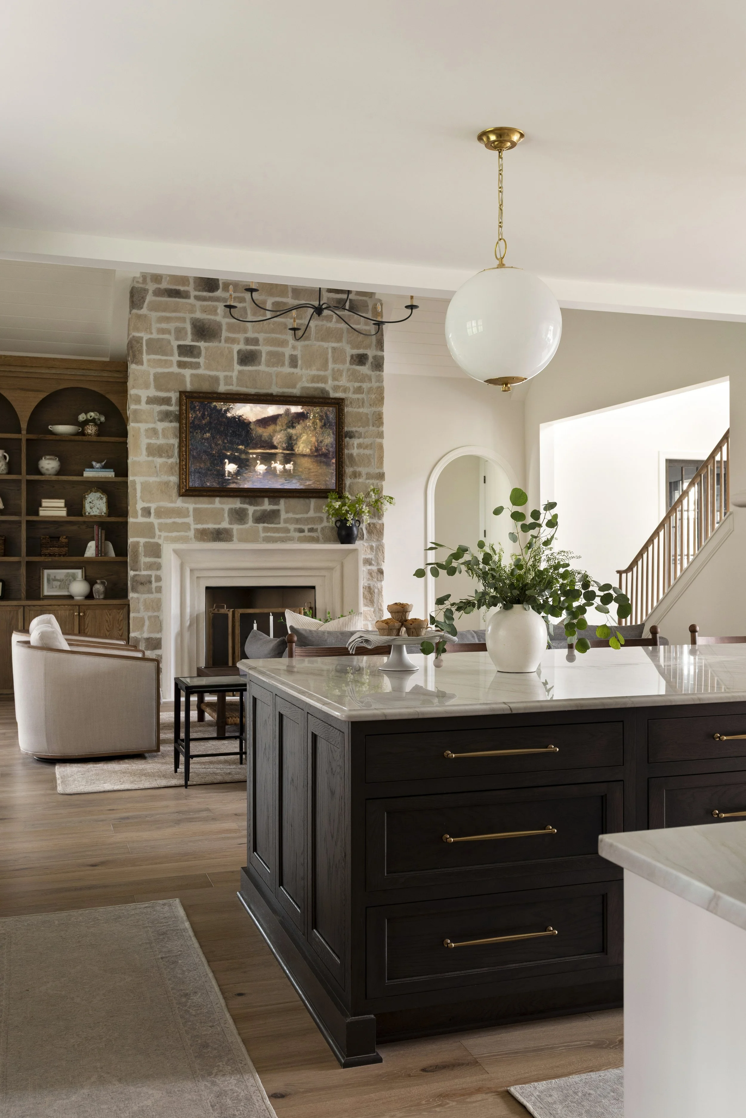 A living room with a brick fireplace, a wooden built-in bookshelf, a beige armchair, wooden side table, and a kitchen island with a white marble top and a large white vase with green foliage.
