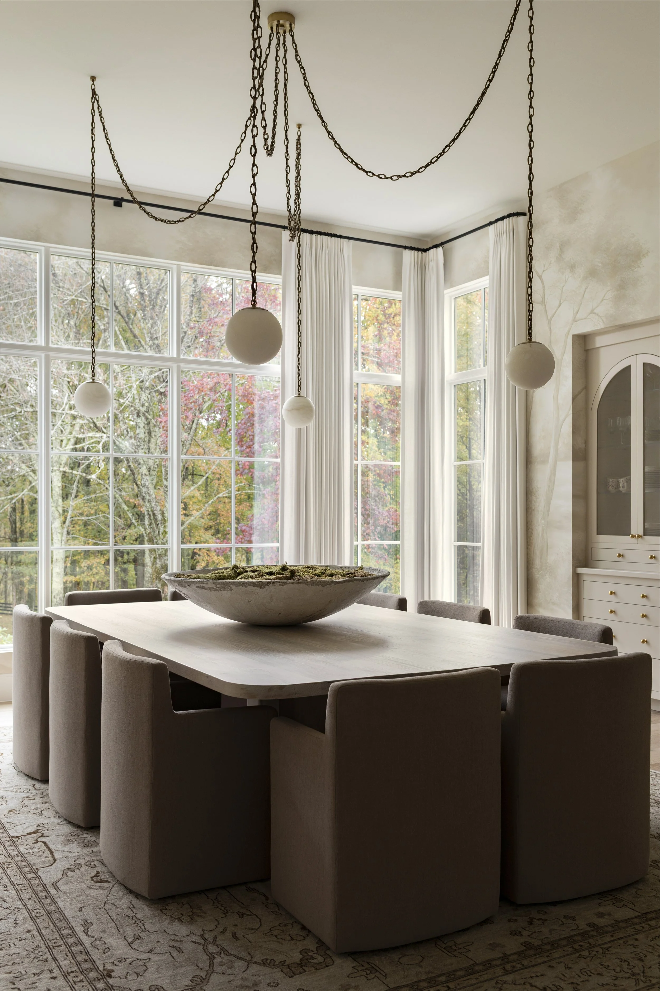 A dining room with a large rectangular wooden table surrounded by twelve beige chairs. A large decorative bowl is on the table. Four hanging light fixtures with globe bulbs hang from chains from the ceiling. Large windows with white curtains show tre