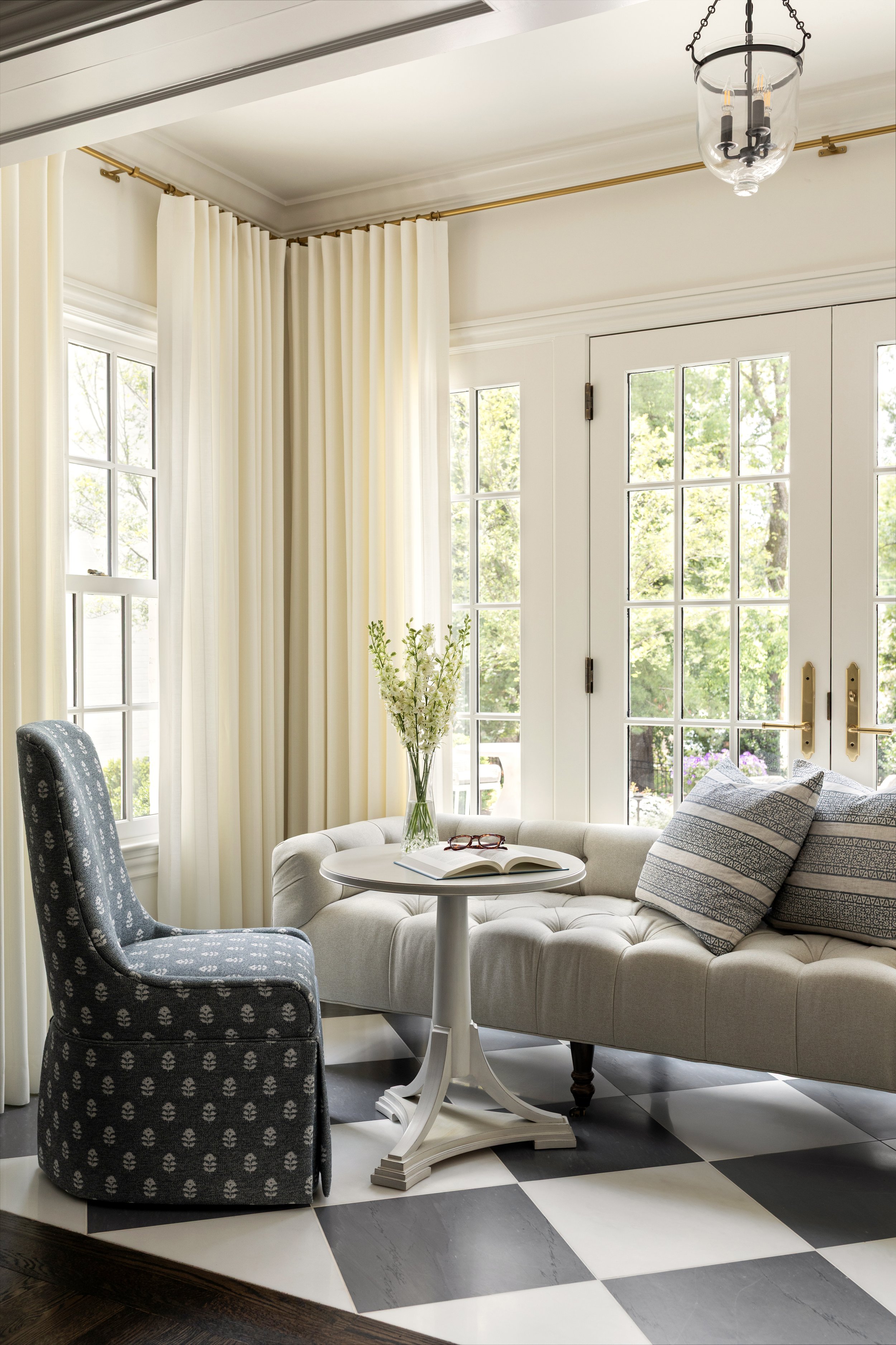 Bright living room with black and white checkered floor, white sofa with striped pillows, dark patterned armchair, small round table with a vase of flowers and reading glasses, behind glass-paneled doors and windows with cream curtains, and a modern ceiling light fixture.