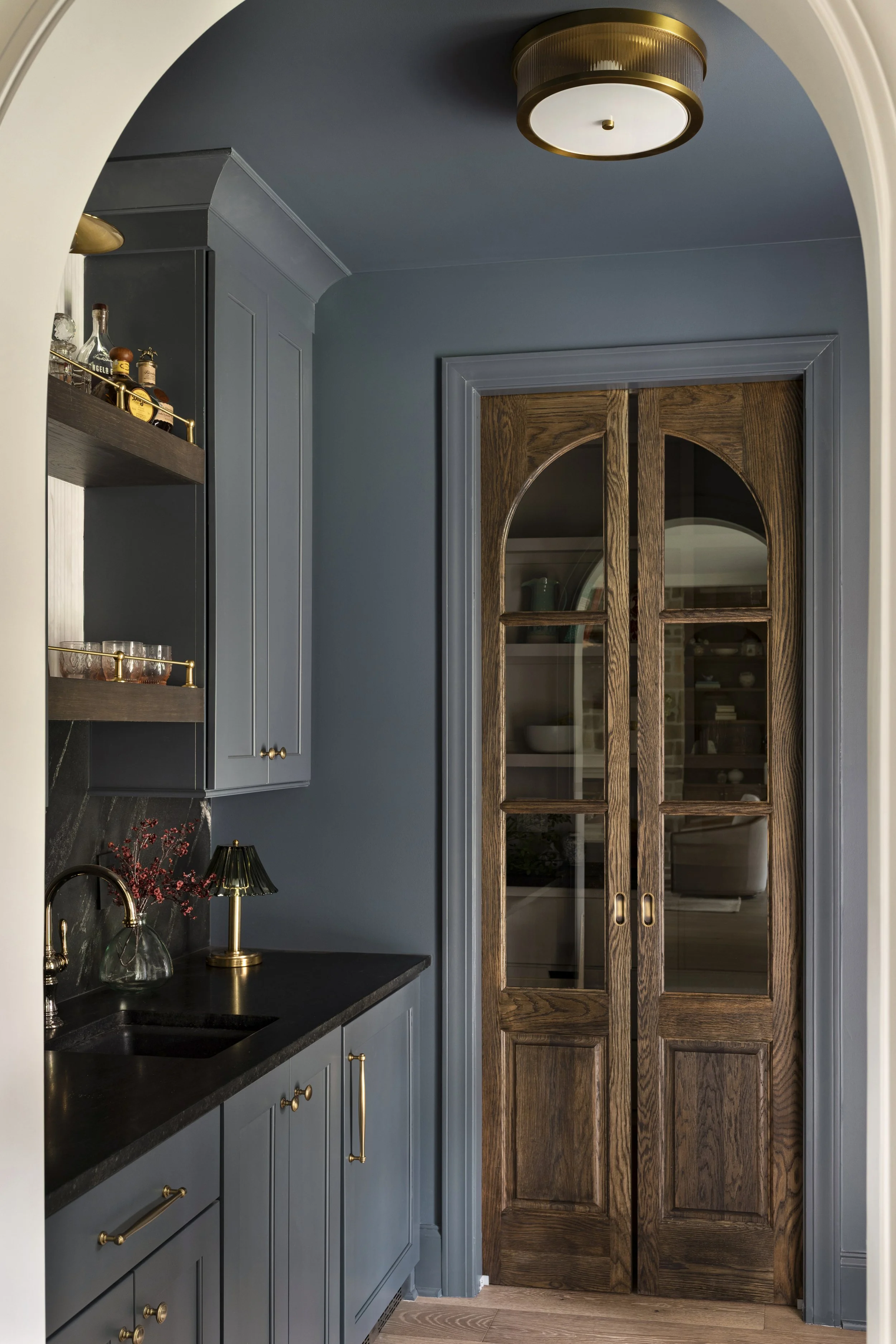 Interior view of a kitchen with blue cabinetry, black countertop, gold hardware, a small black and gold lamp, a clear glass vase with red berries, and wooden arched double doors with glass panes.