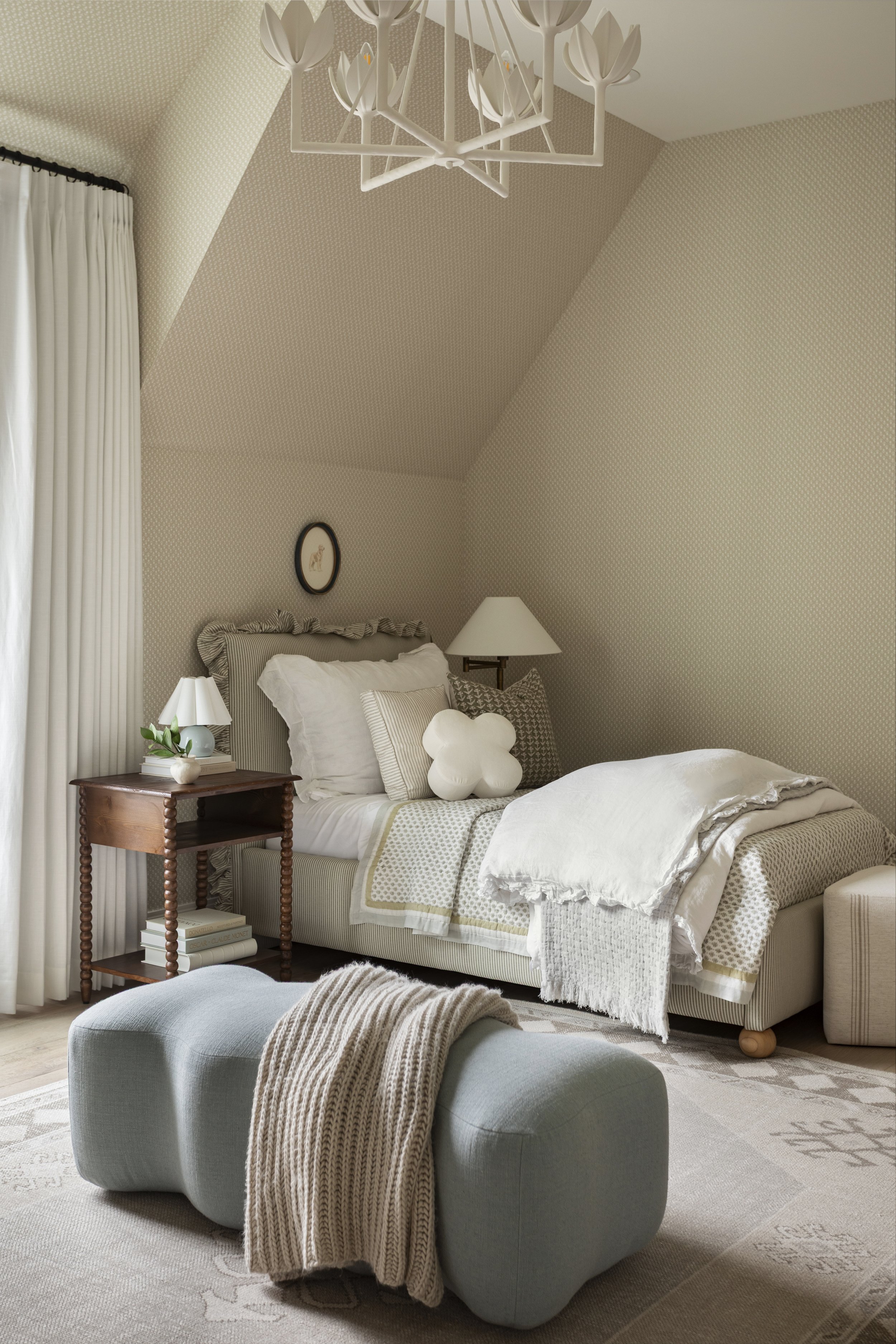 A cozy bedroom with a sloped ceiling, cream and beige decor, a bed with white bedding and decorative pillows, a wooden nightstand with a table lamp and books, a light blue upholstered ottoman with a beige knitted throw, and white curtains.