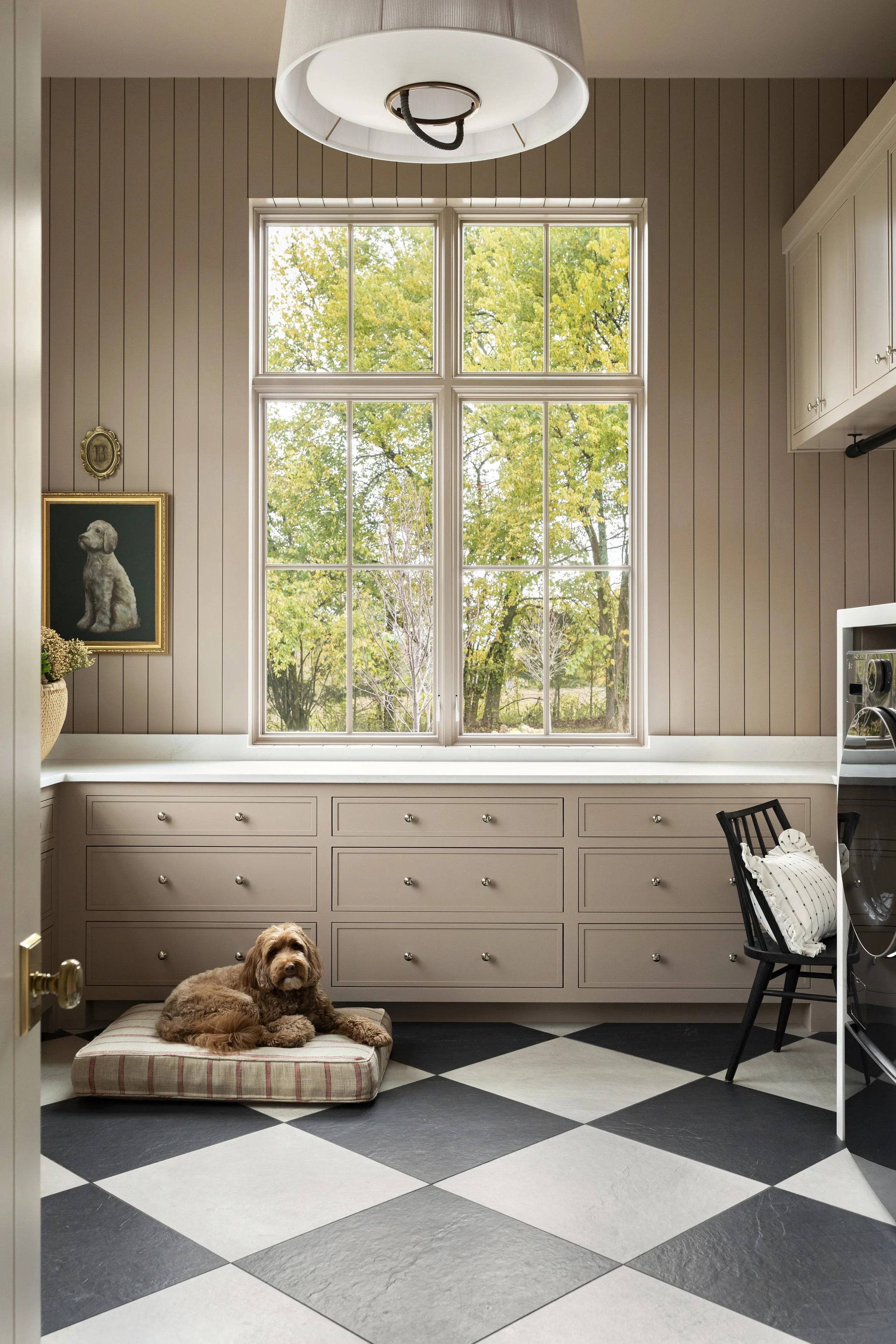 A cozy room with a large six-pane window showing green trees outside, beige wood-paneled walls, a plaid dog bed with a brown dog, black and white checkered tile flooring, a black chair with white pillows, and a laundry appliance.