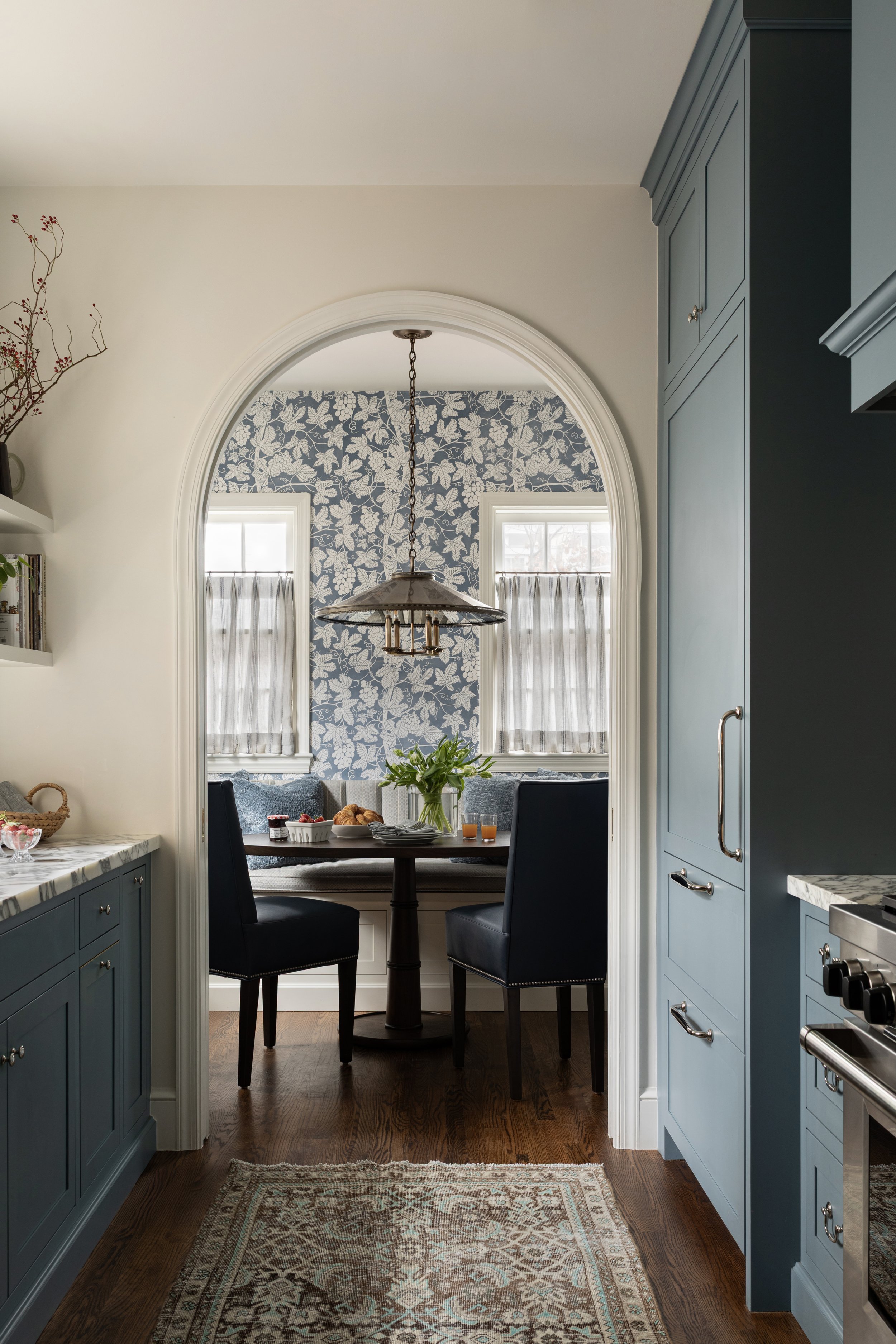 View through a doorway into a cozy dining nook with a round table, four black chairs, a floral arrangement, and two glasses of what appears to be orange juice. The area has two windows with sheer curtains and features blue-blue patterned wallpaper wi