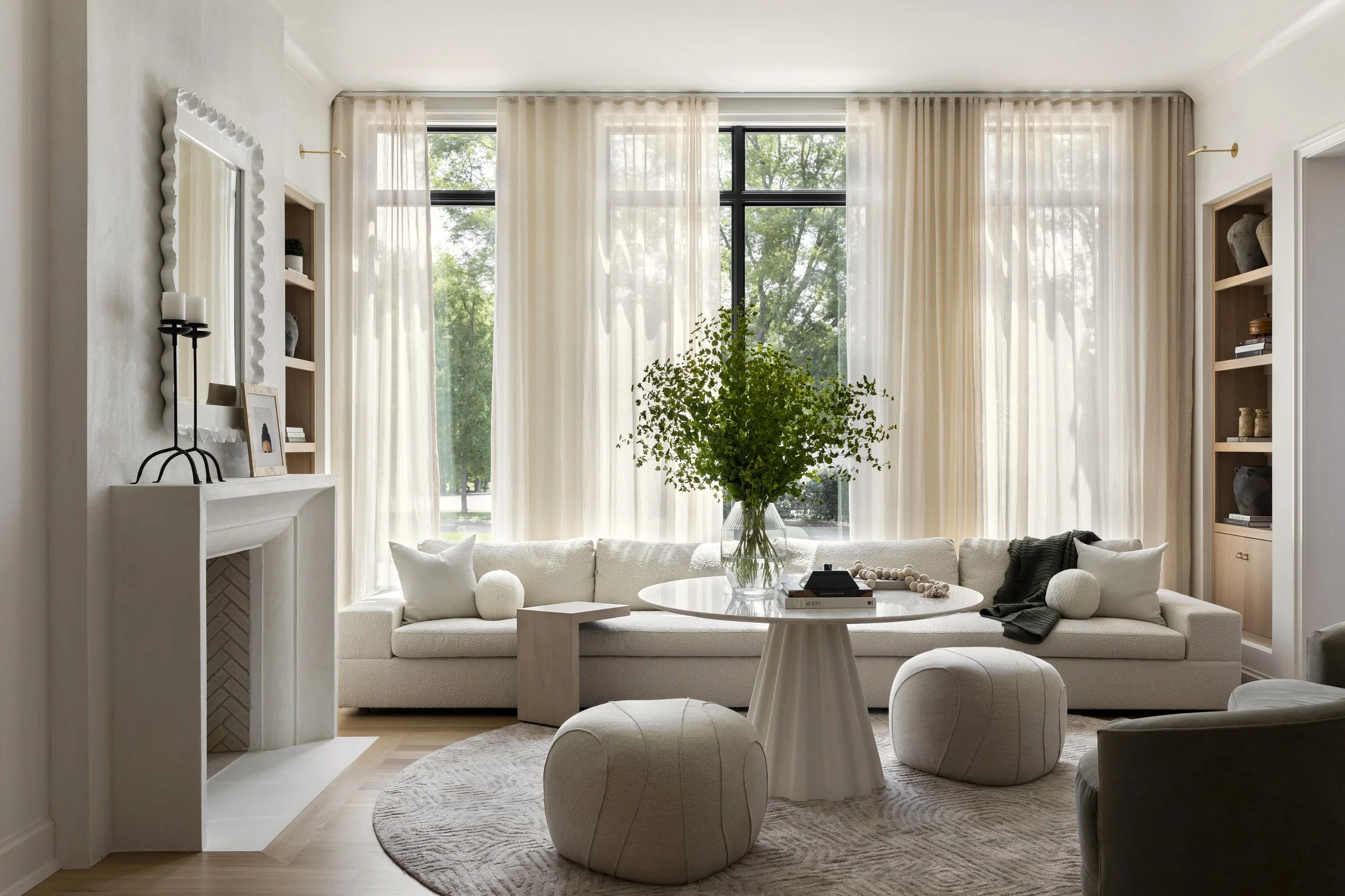 Living room with cream-colored sofa, round coffee table with a large plant, two matching poufs, and beige curtains covering tall windows with trees outside.