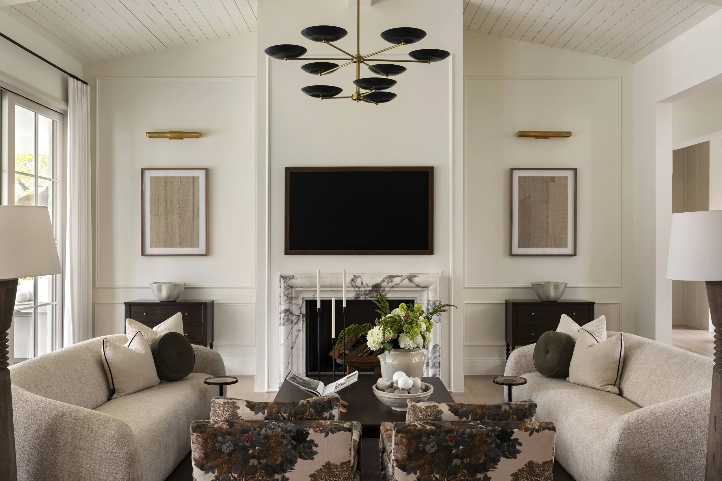 Living room with two beige sofas, floral armchairs, a black coffee table with flowers, a fireplace, two side tables with lamps, wall art, and a large wall-mounted TV.