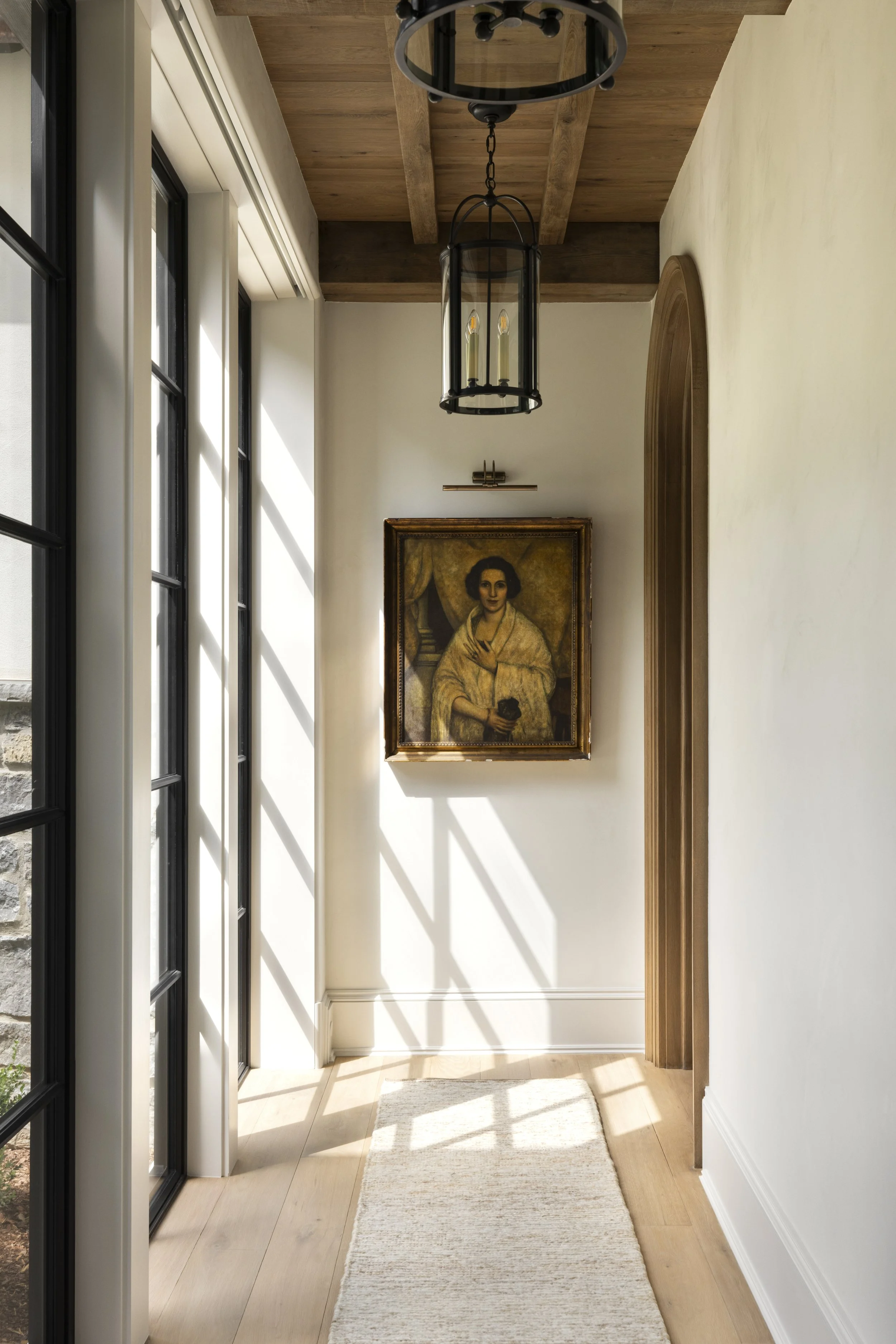 Sunlit hallway with large windows, a hanging lantern light fixture, a framed portrait of a woman on the wall, and a white rug on a wooden floor.