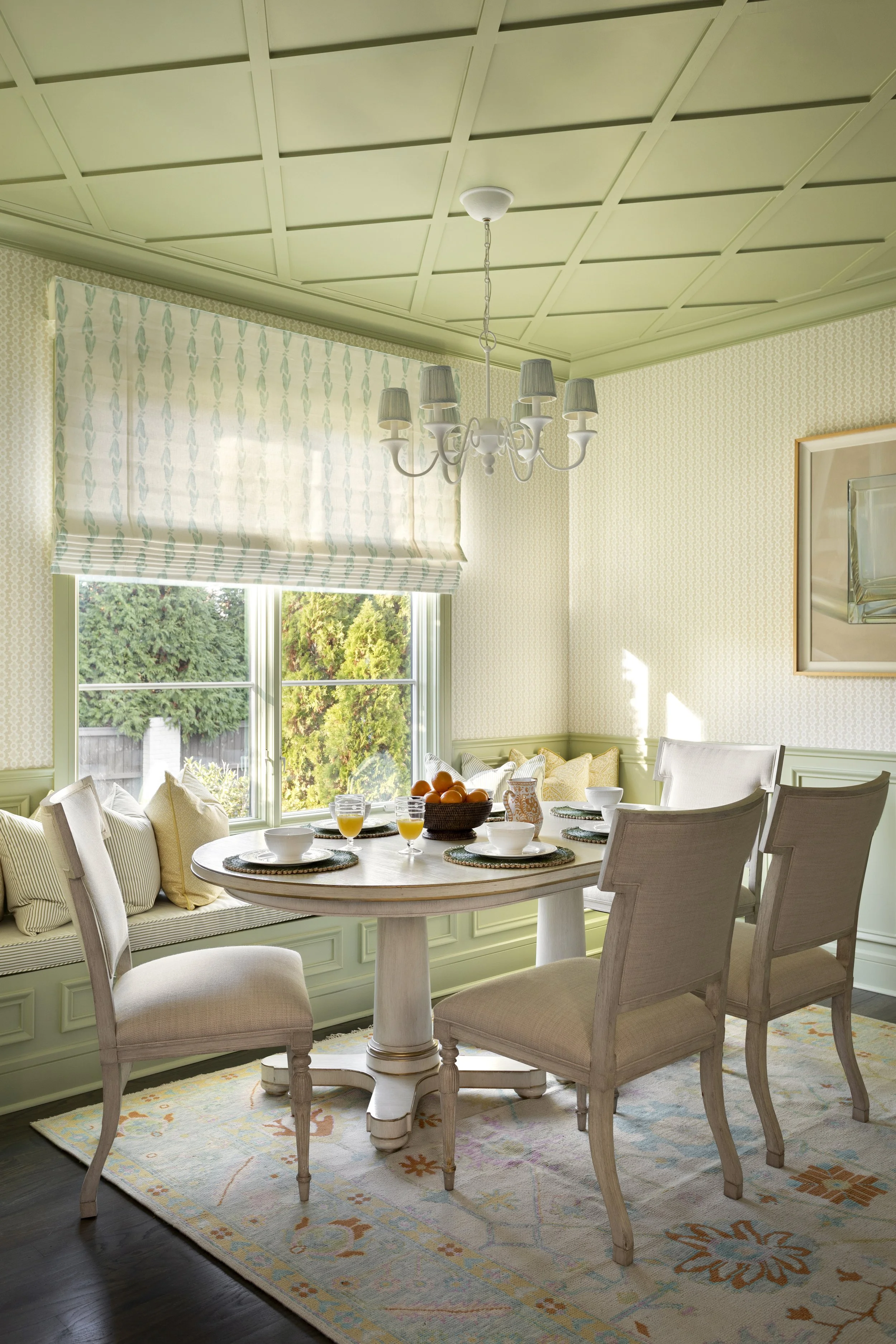 A dining room with light green walls and ceiling, a window with a striped Roman shade, a round wooden table set for breakfast with orange juice, bowls, and a basket of oranges, surrounded by upholstered chairs, a patterned area rug, and a chandelier 