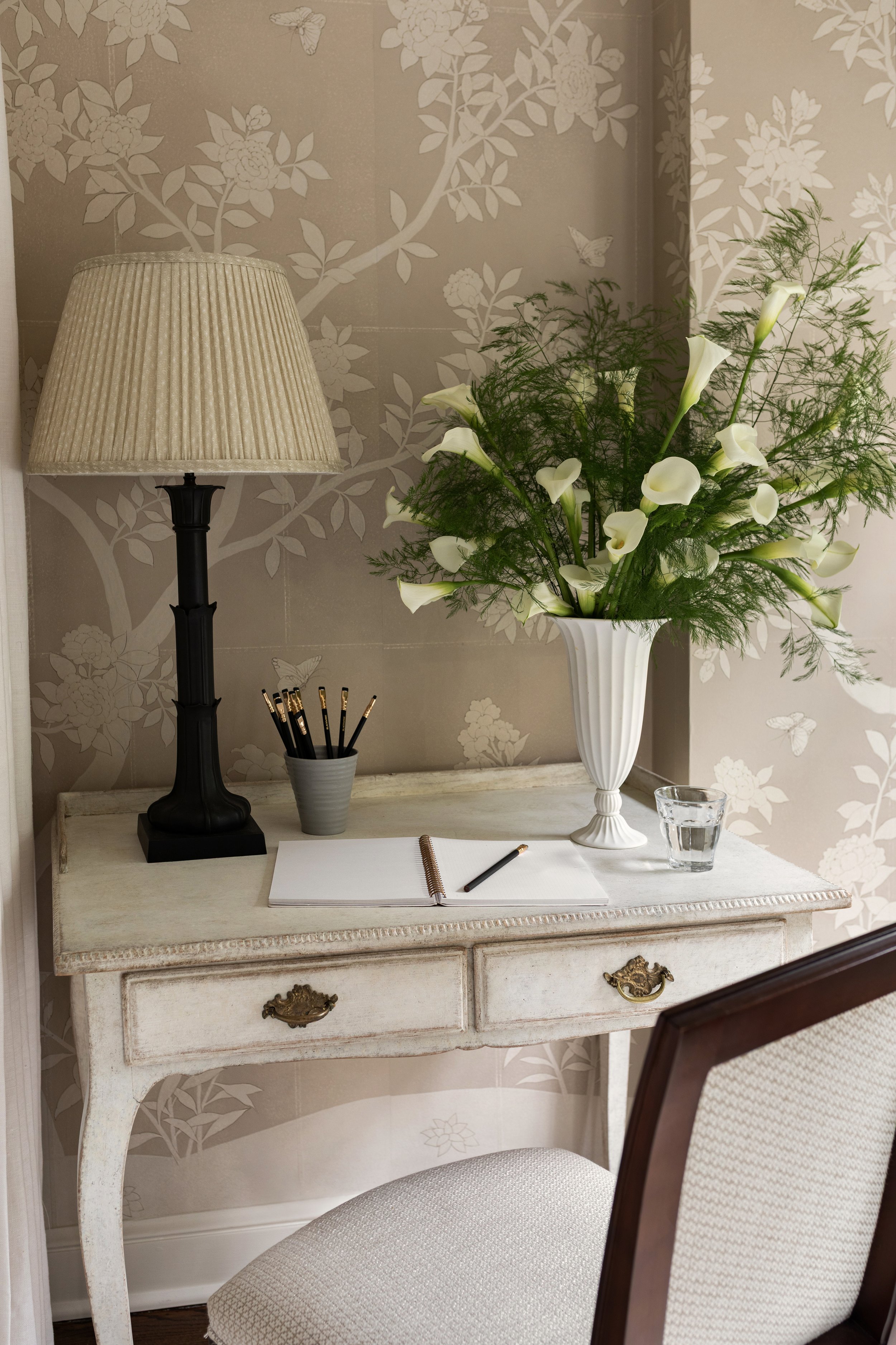 A vintage white desk with a floral design, a beige table lamp, a gray cup with black and gold pencils, an open notebook with a pen, a glass of water, and a white vase with white calla lilies and green foliage, set against a wallpapered wall with butterfly and floral patterns.