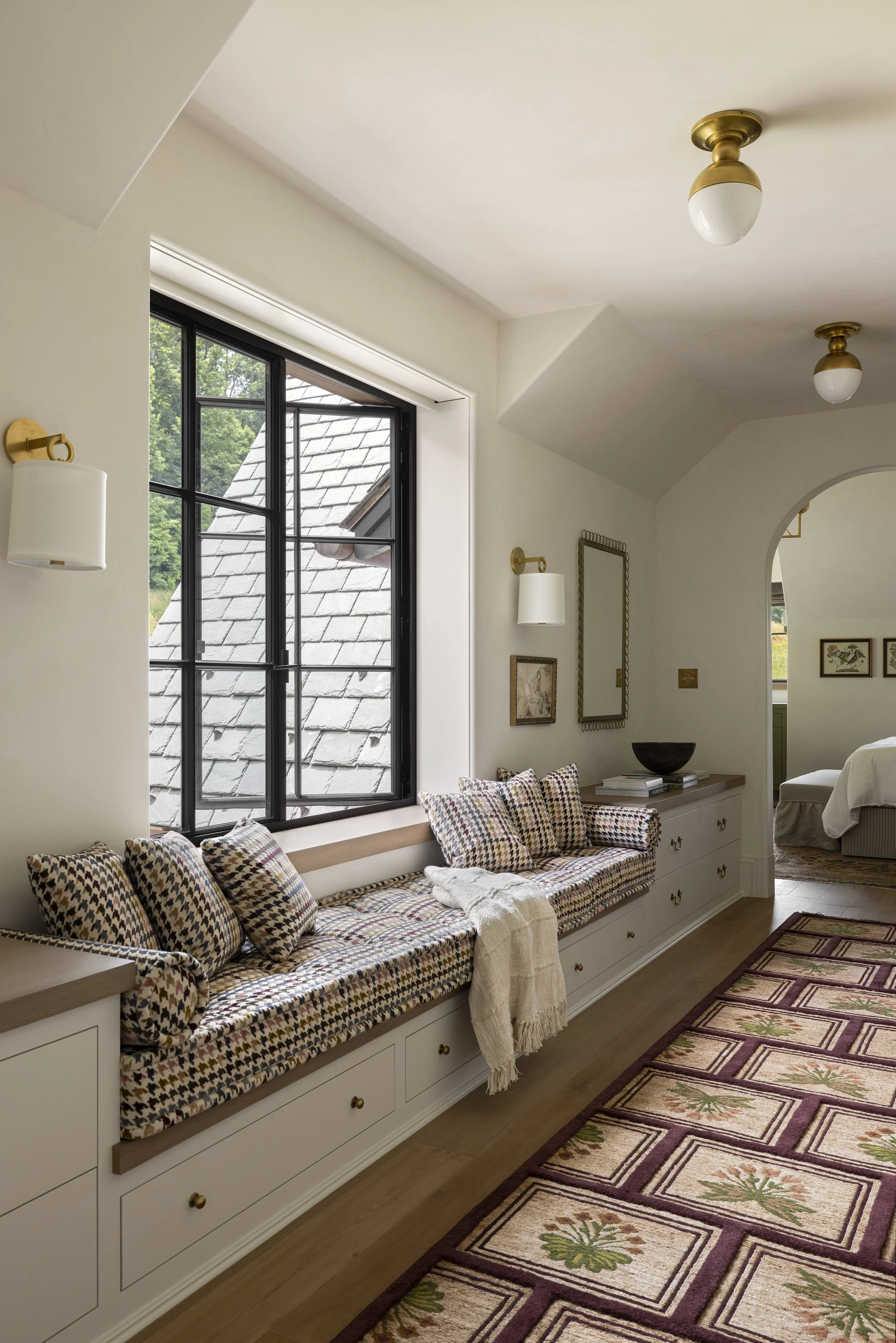 A window seat with patterned cushions and a throw blanket, sunlight coming through a large window, with a view of a roof and trees outside.