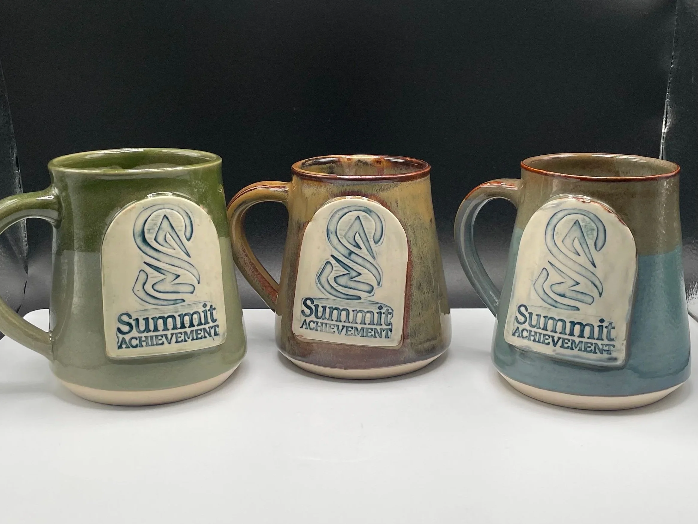 A ceramic jar with a blue lid sitting outdoors on a rocky surface in a desert landscape with dry grasses, rocks, and mountains in the background under a clear blue sky.