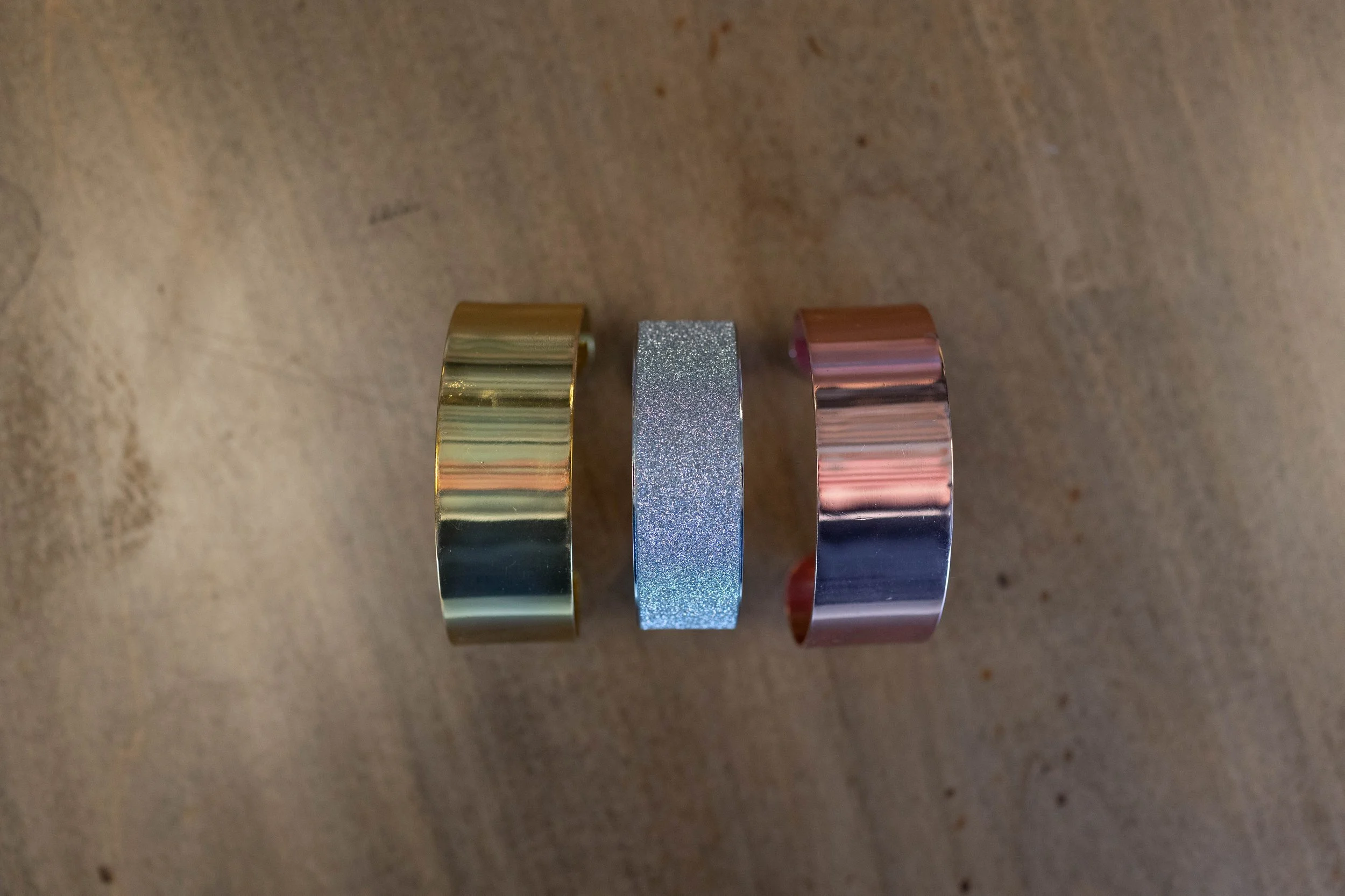 Three metallic rings placed on a wooden surface, from left to right: gold-colored, silver glittery, and rose gold-colored.