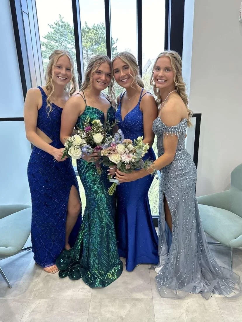 Four young women in formal dresses posing together, holding bouquets, inside a room with large windows showing trees outside.