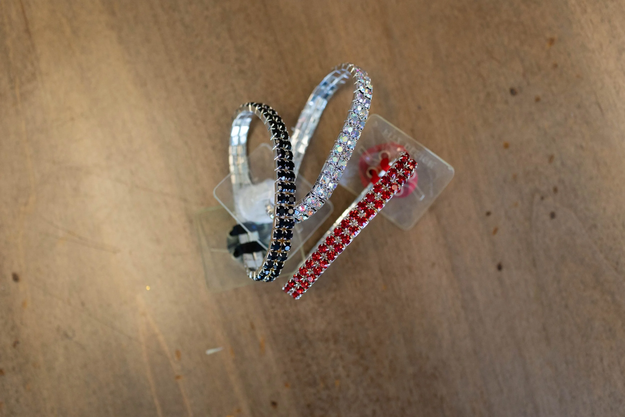 Three rhinestone headbands in black, silver, and red, displayed on a transparent plastic stand on a wooden surface.