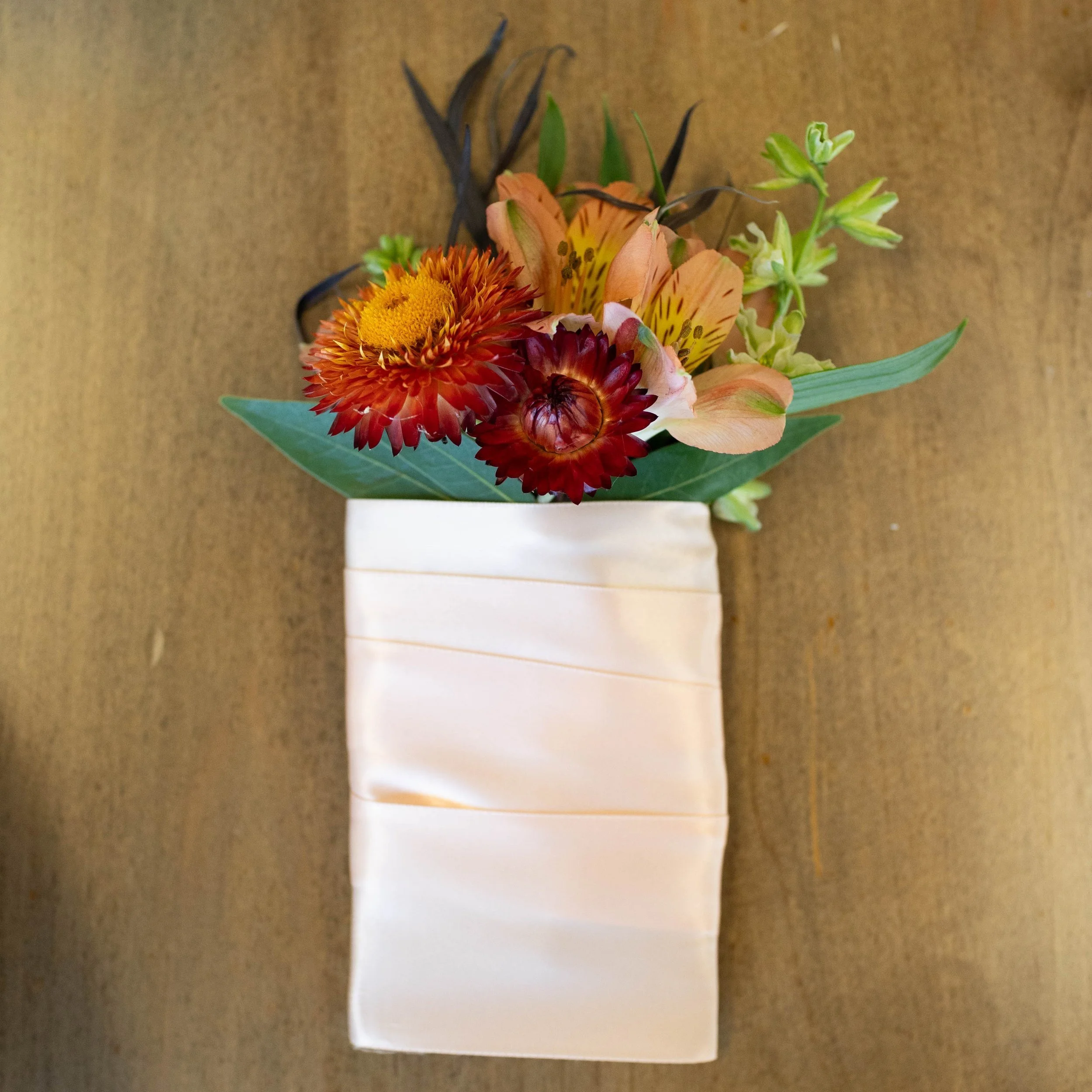 A bouquet of orange, red, and yellow flowers in a cream-colored, textured vase on a wooden surface.