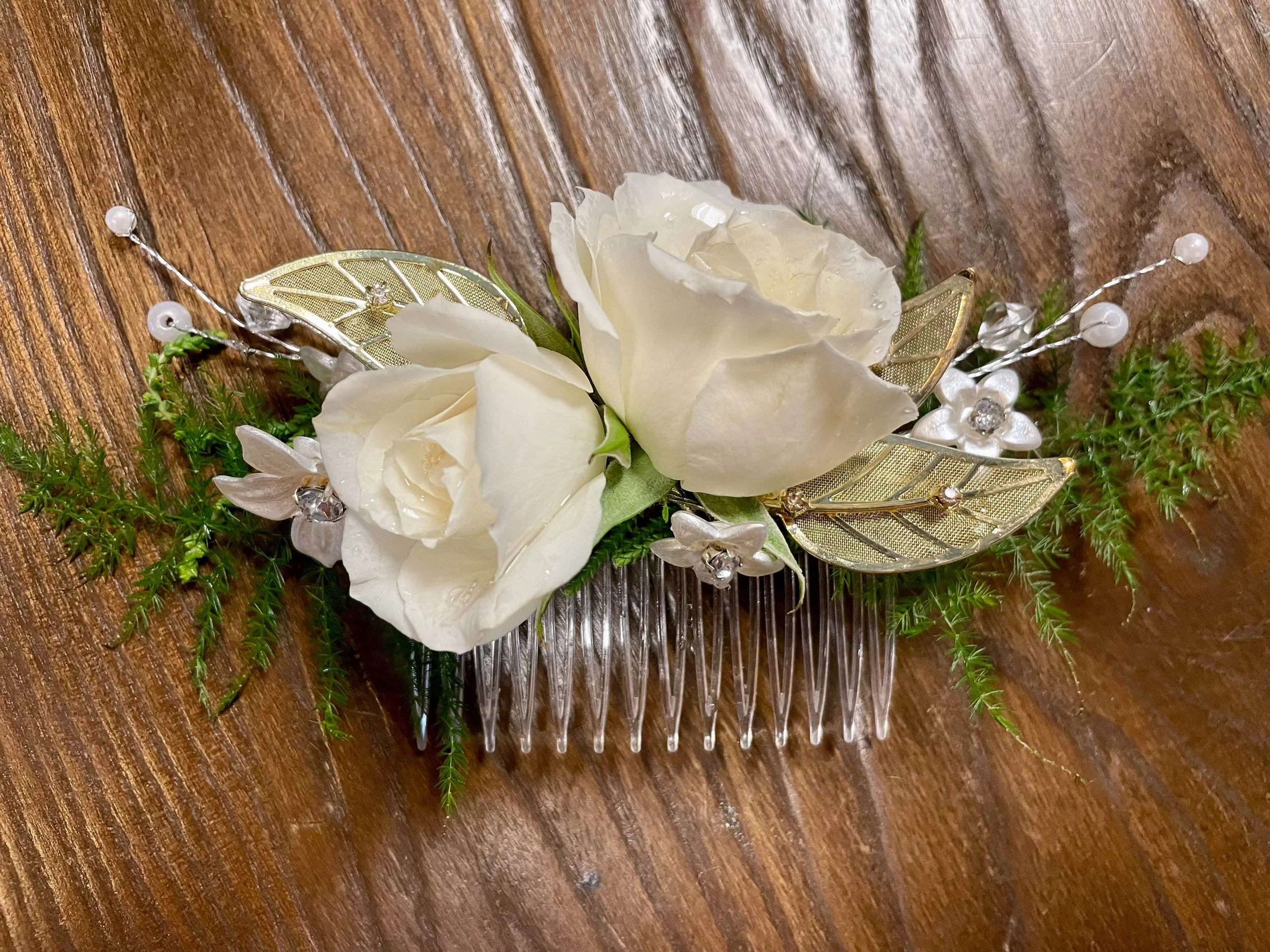 A decorative hair comb with white roses, gold butterfly wings, green foliage, small white flowers, and clear rhinestones on a wooden surface.